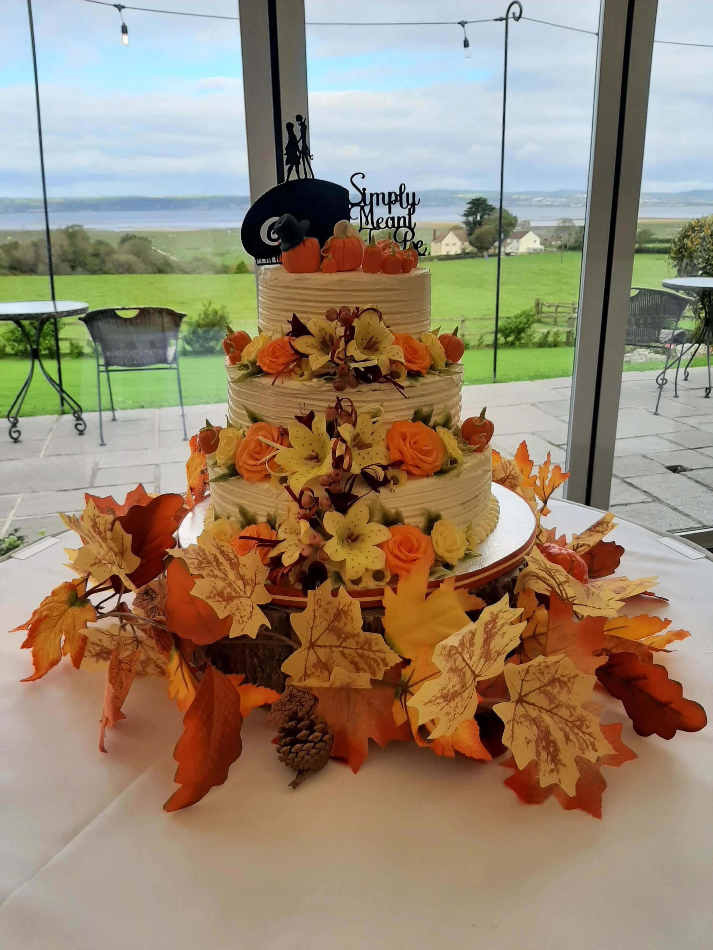 Three-tiered wedding cake decorated with orange roses, yellow lilies, autumn leaves, pinecones, and small pumpkin toppers, set on a tree stump surrounded by fall leaves and pinecones, with a scenic landscape view through large windows.