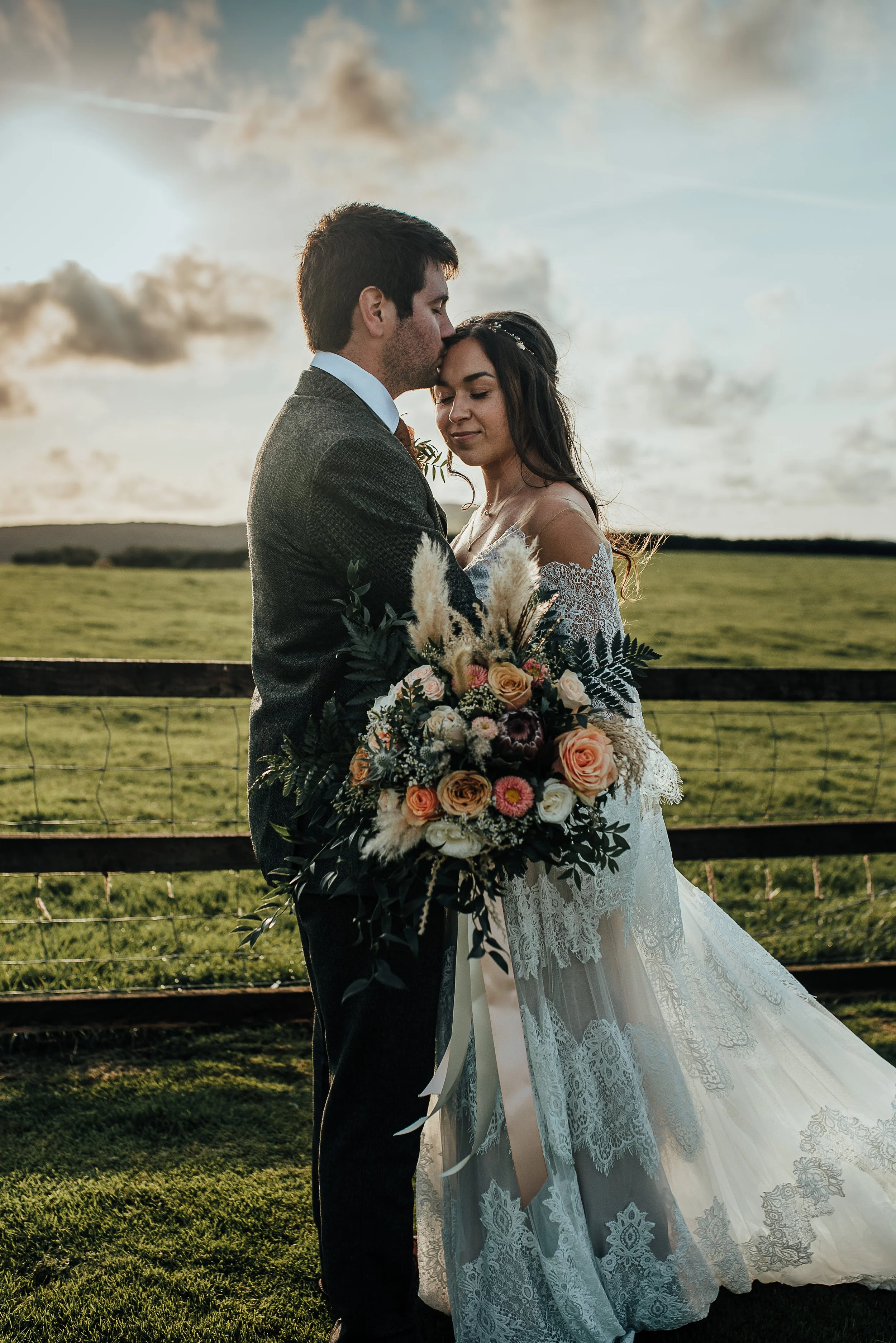 A couple in wedding attire standing close together outdoors, with the man kissing the woman's forehead while holding a large bouquet of flowers. The scene is set in a grassy field during sunset.