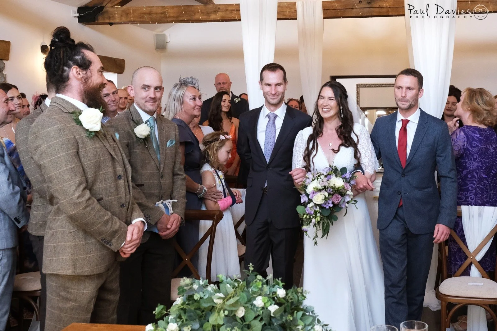 Wedding ceremony with the bride and groom standing in the center, surrounded by family and friends in a decorated indoor venue.