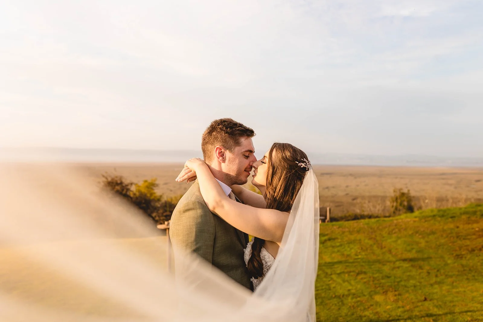 Bride and groom embracing on a grassy field with a veil flowing in the foreground.