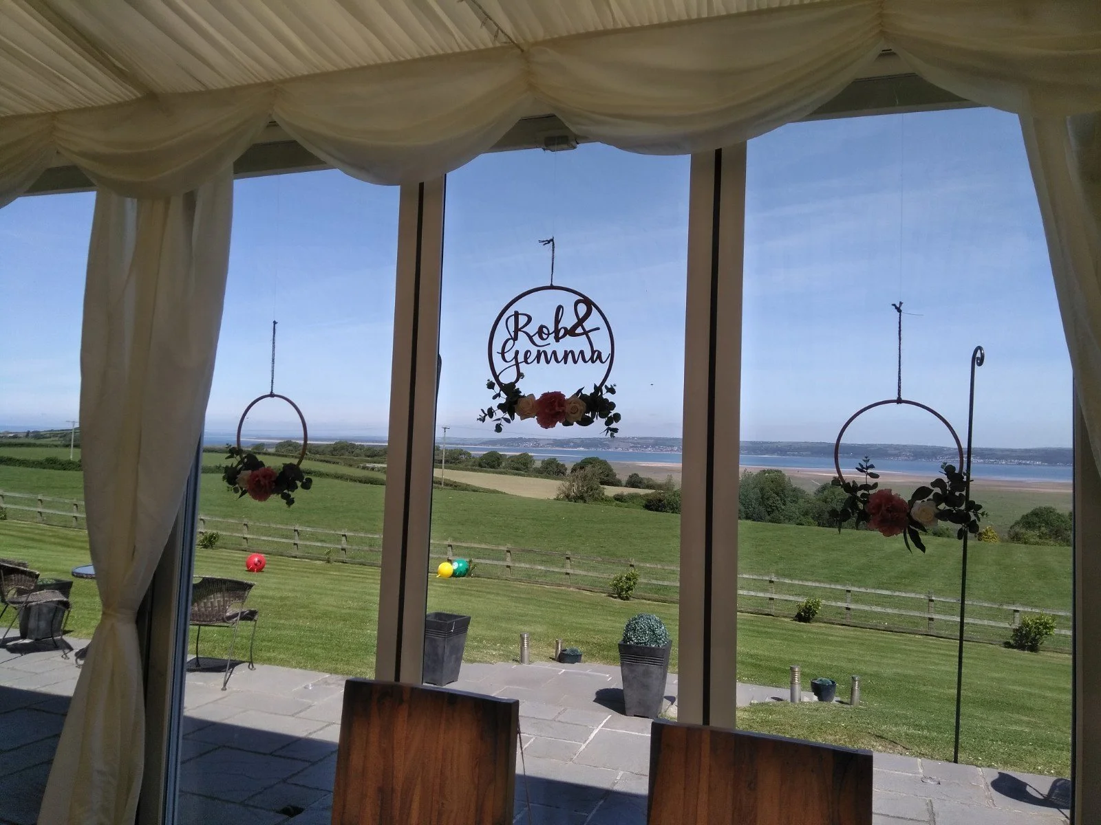 Decorative hanging floral signs with the text 'Rob & Gemma' inside a circular frame against a view of a grassy outdoor landscape with a river and blue sky.