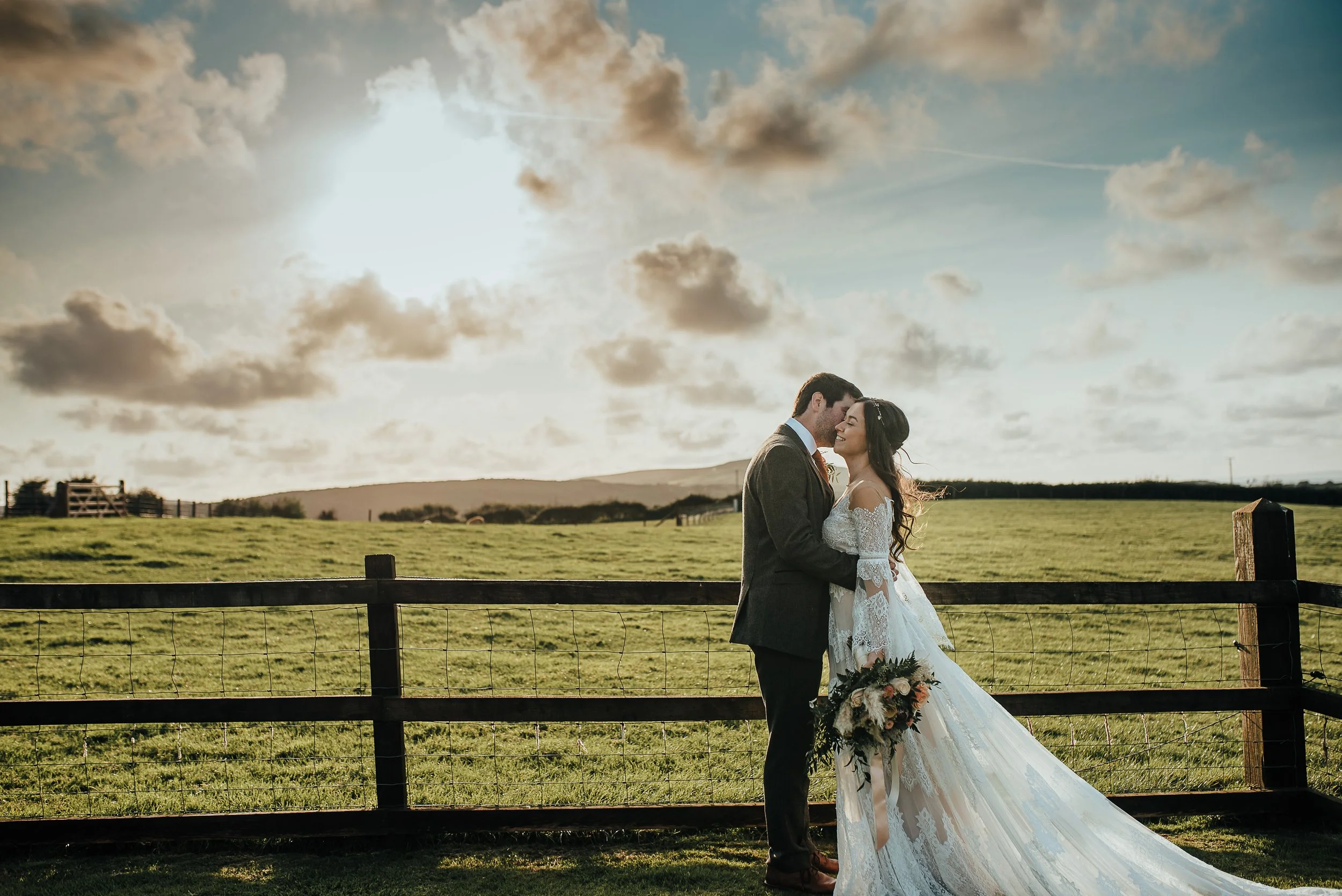 A bride and groom sharing a kiss outdoors on their wedding day, with a green field, a wooden fence, and a cloudy sky at sunset in the background.