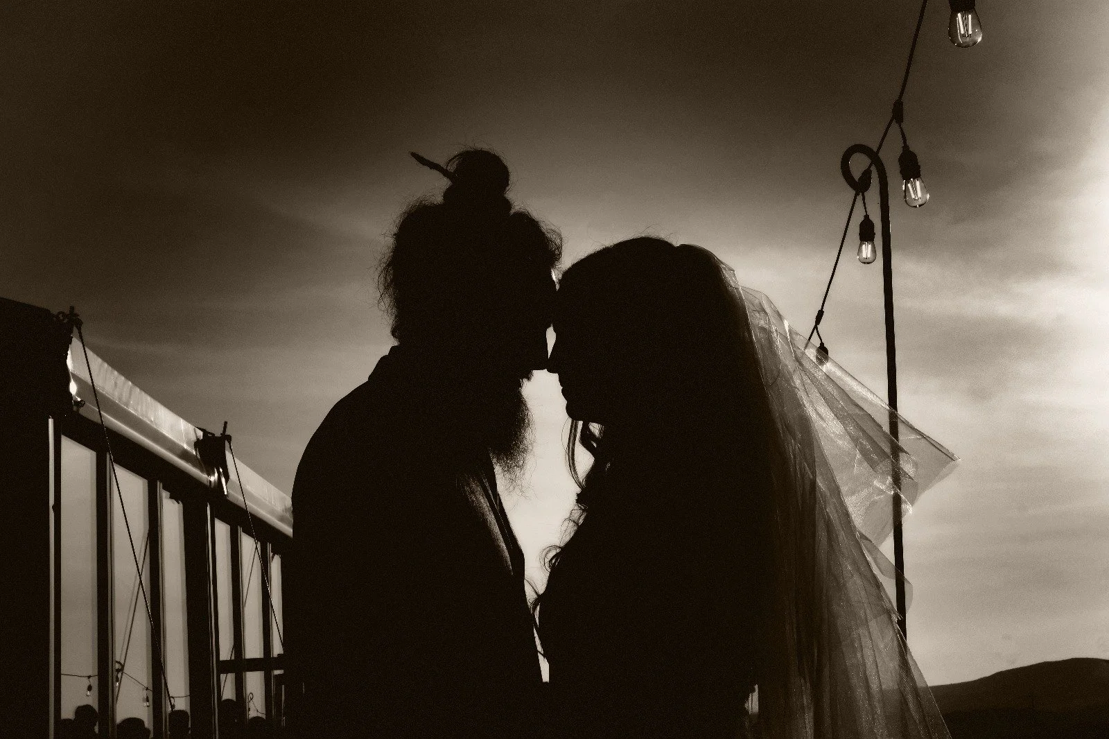 Silhouette of a couple with noses touching, with a bearded man in a bun and a woman with long hair and a veil, outdoors during sunset with string lights hanging overhead.
