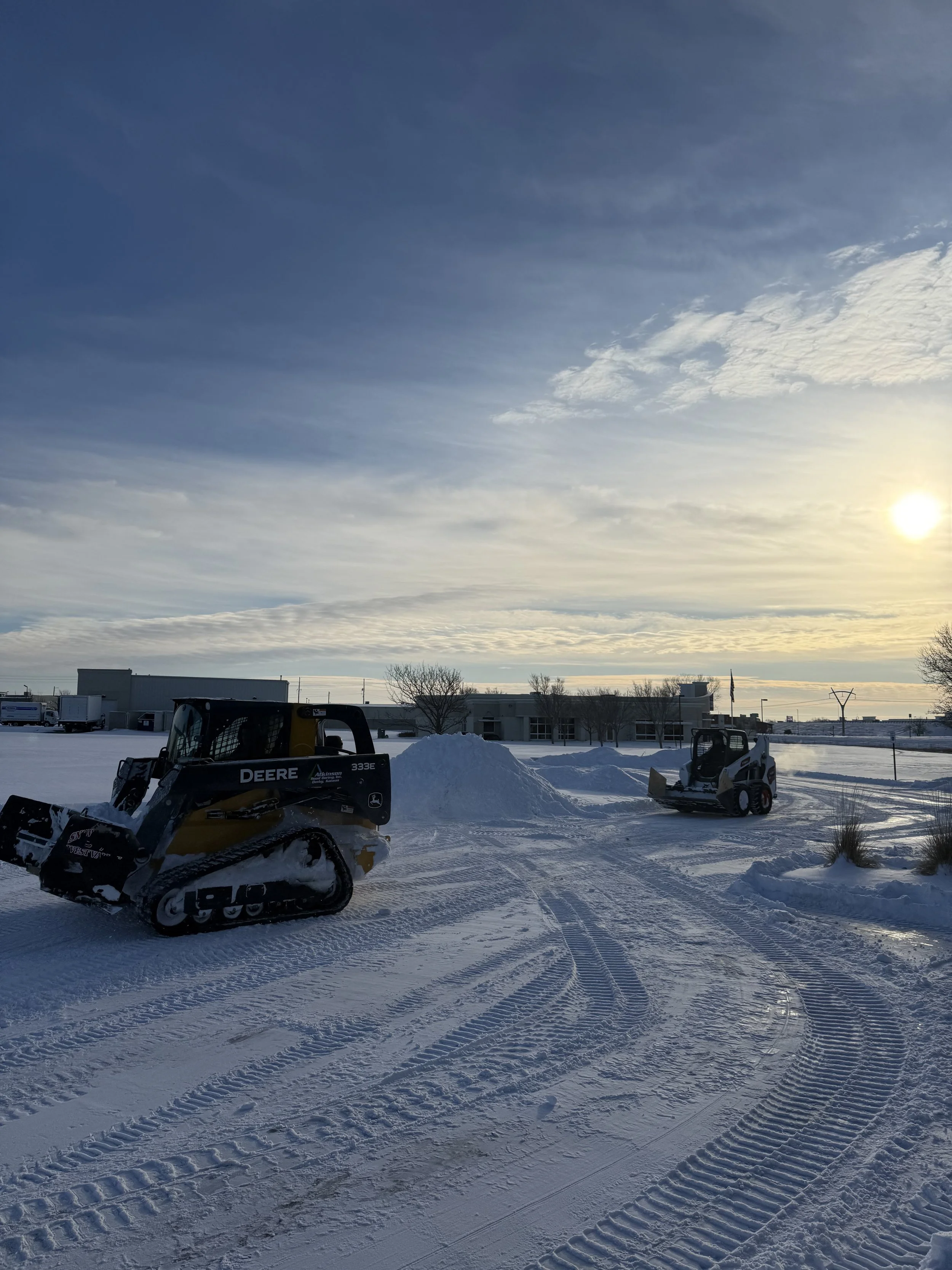 Snow-covered parking lot with two skid-steer loaders and piled snow, under a cloudy sky with the sun setting.
