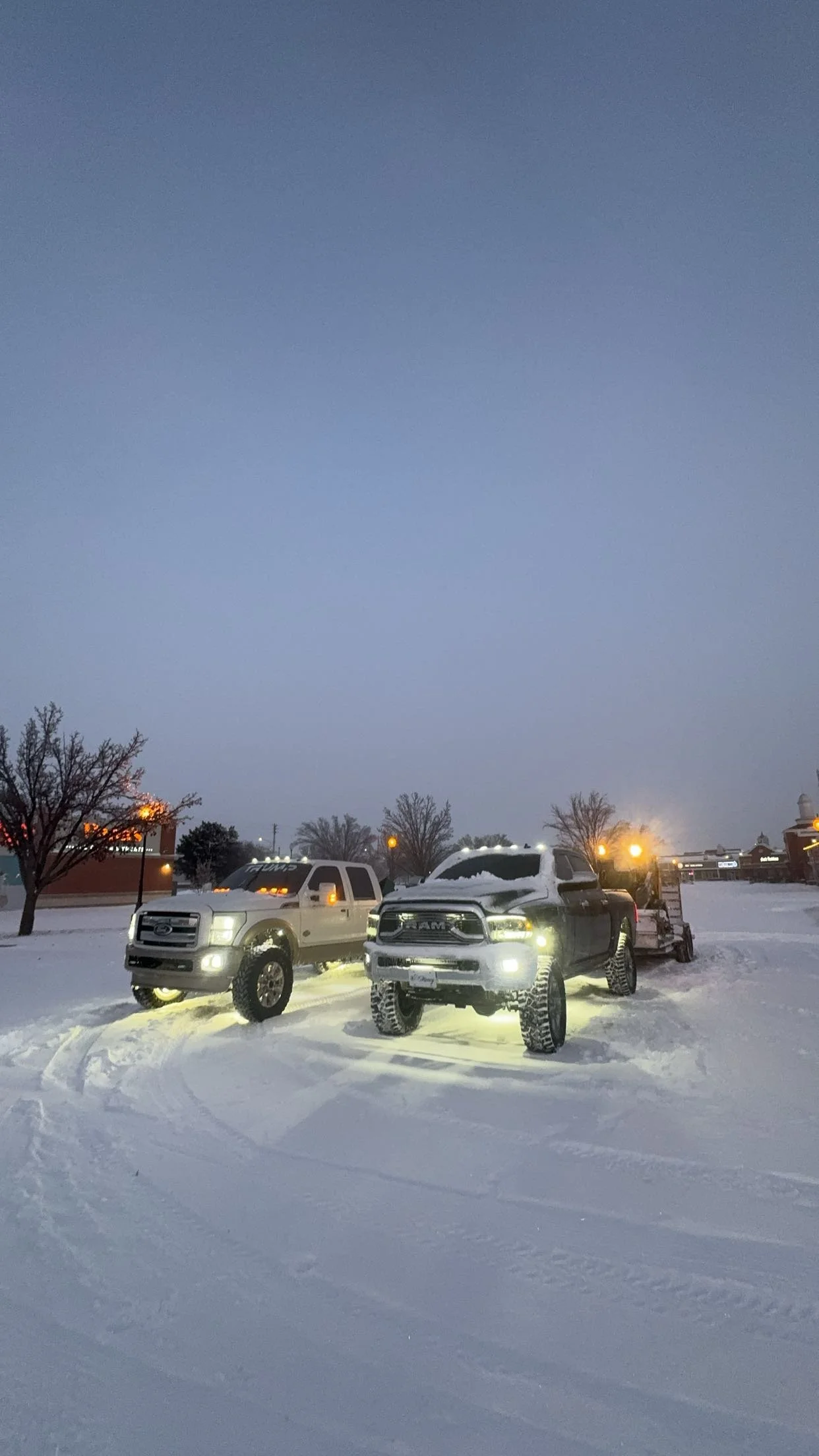 Two pickup trucks parked on snow-covered ground at dusk, with snow on the vehicles and a trailer hitched to one of the trucks. There are trees with holiday lights and some buildings in the background.