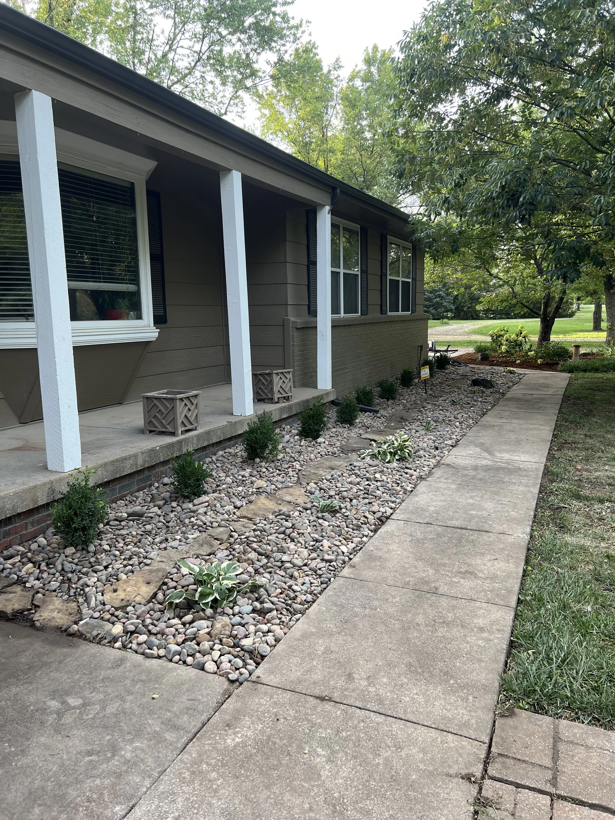 Front yard and porch of a house with a stone and pebble garden bed, small shrubs, a sidewalk, and trees in the background.