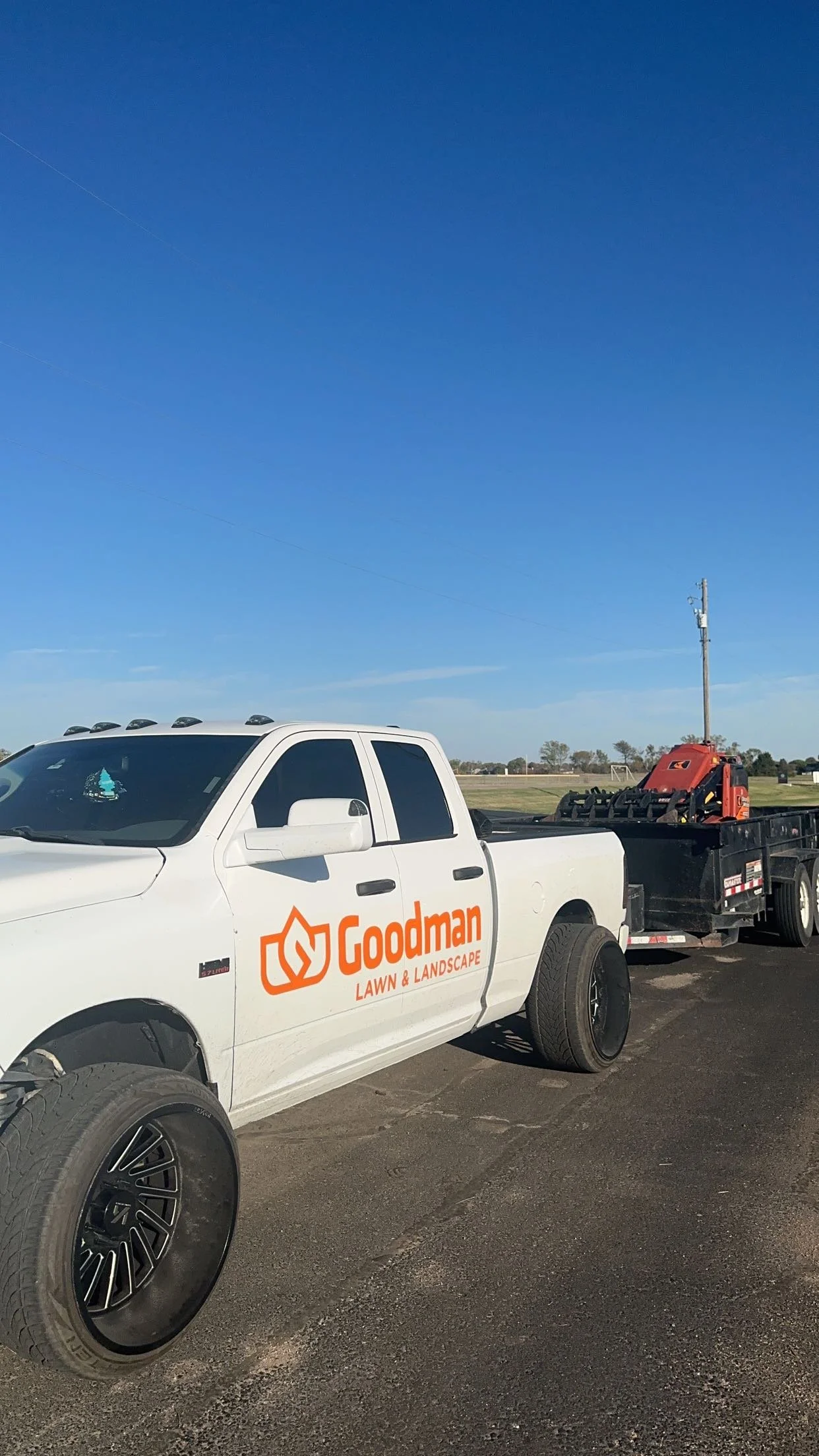 A white pickup truck with the logo and name 'Goodman Lawn & Landscape' parked on a paved surface, connected to a trailer with equipment, under a clear blue sky.