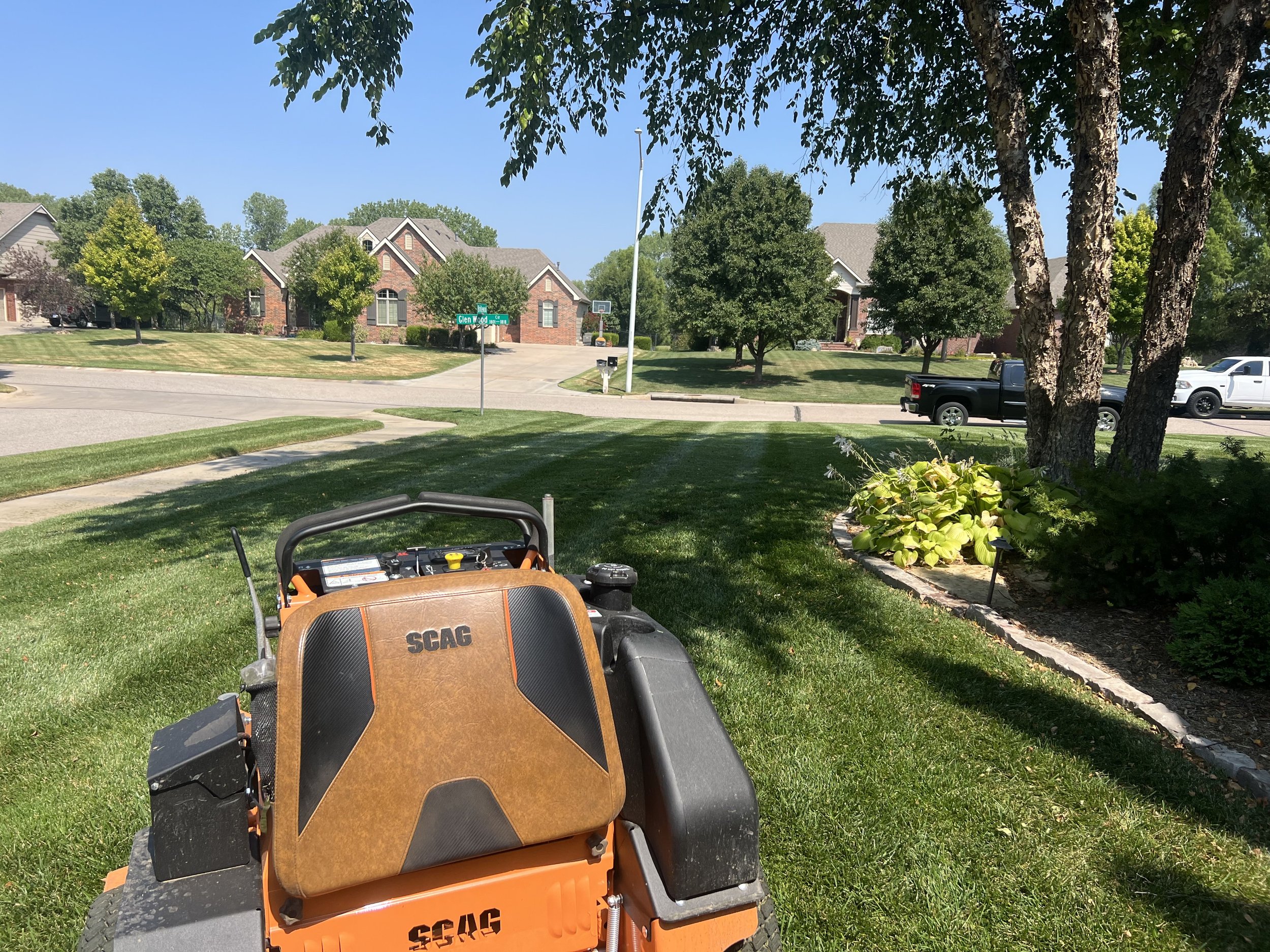 View of a suburban lawn with a riding lawn mower in the foreground, trees, houses, and parked trucks in the background on a sunny day with a clear blue sky.
