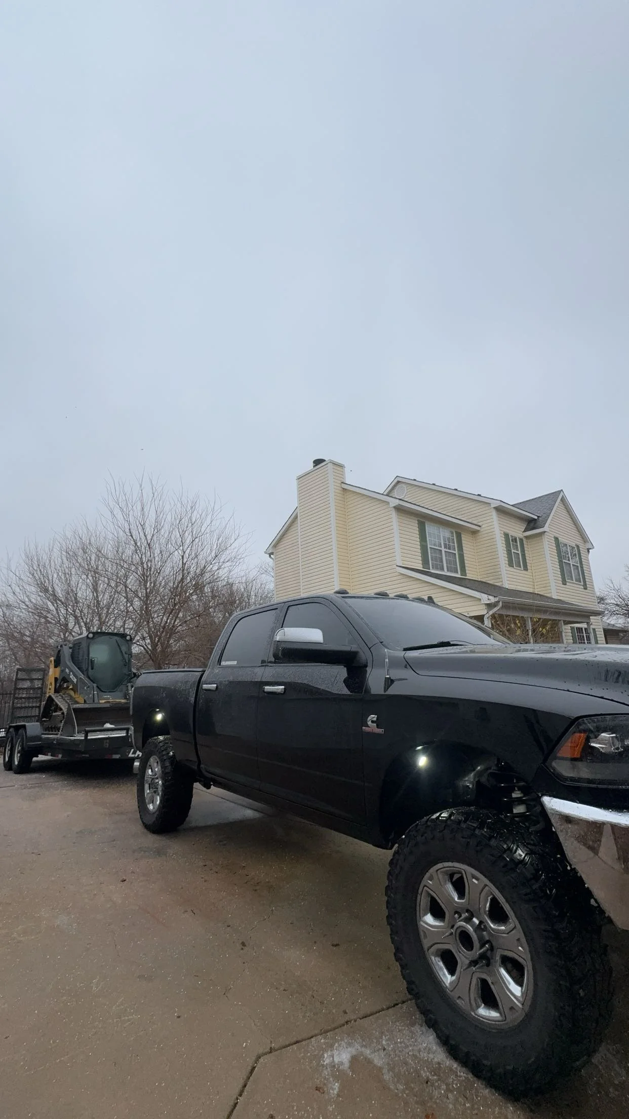 A black pickup truck with large off-road tires parked on a driveway, towing a trailer with a small construction vehicle, in front of a beige two-story house with white trim on a cloudy, overcast day.
