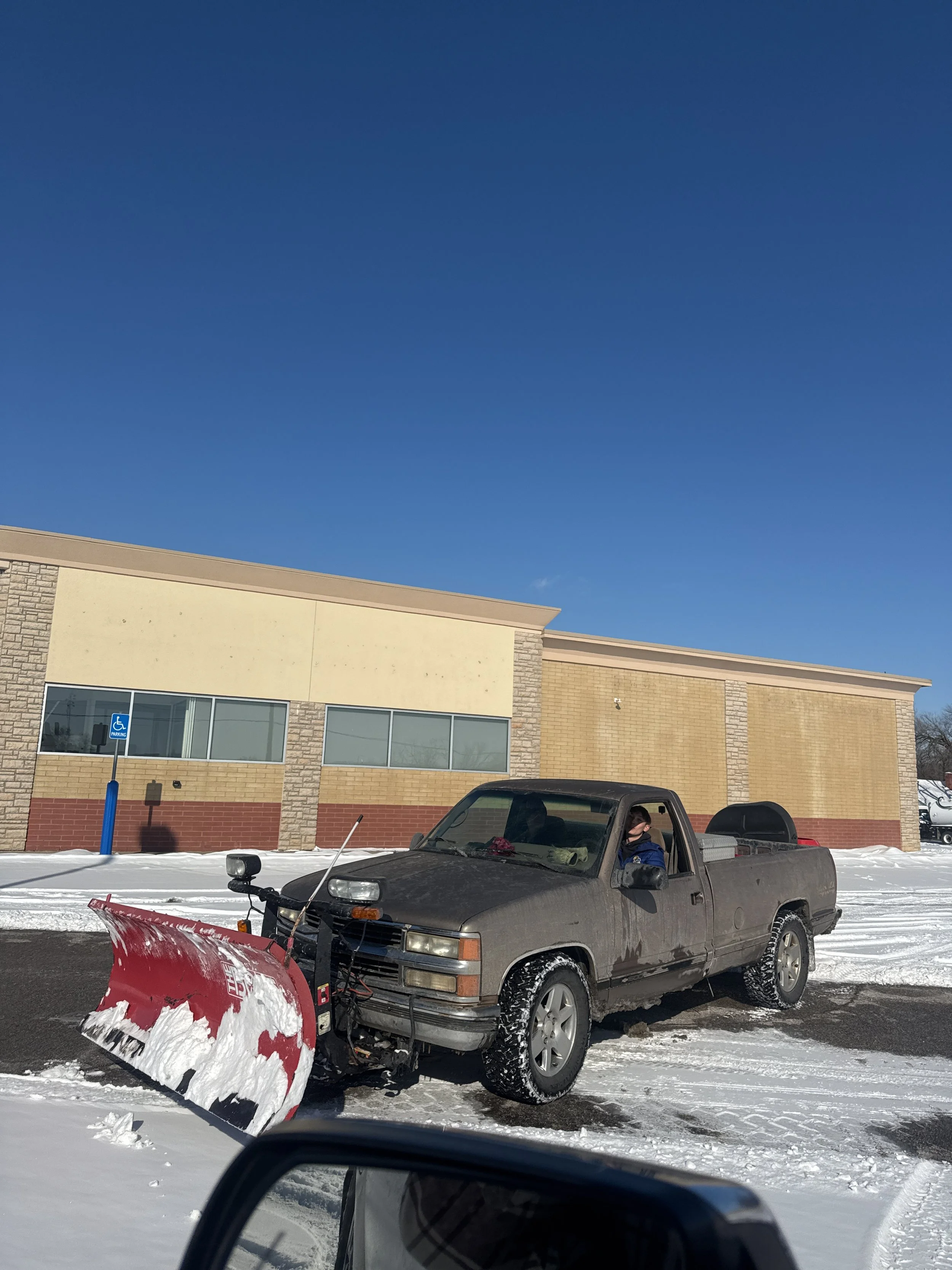 A black pickup truck with a snow plow attached is parked on a snowy and icy parking lot in front of a brick building with large windows. A person wearing a face mask is sitting in the driver's seat.
