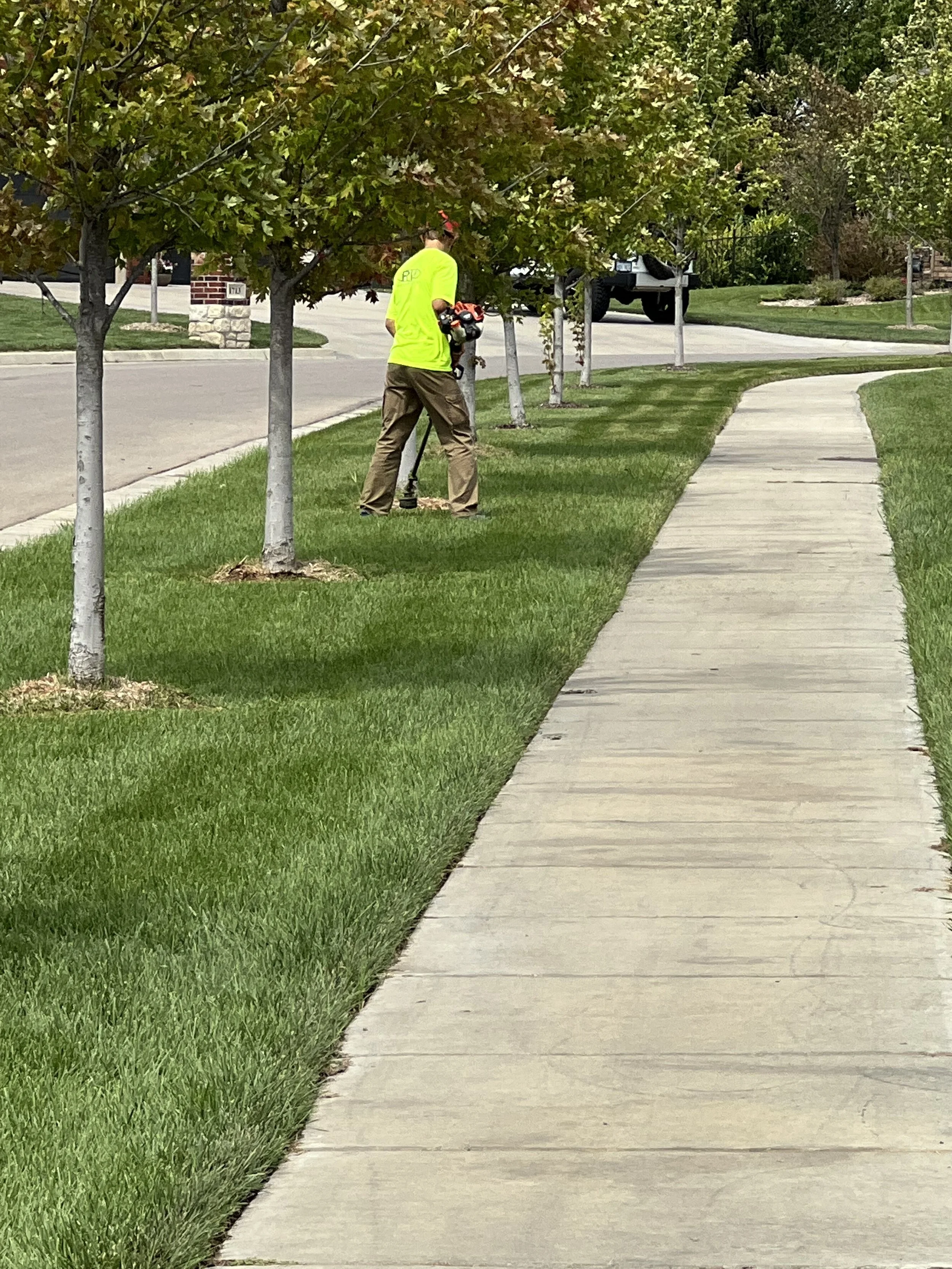 A person wearing a neon yellow shirt and brown pants is using a landscaping tool near the trees on a grassy strip next to a sidewalk in a suburban neighborhood.