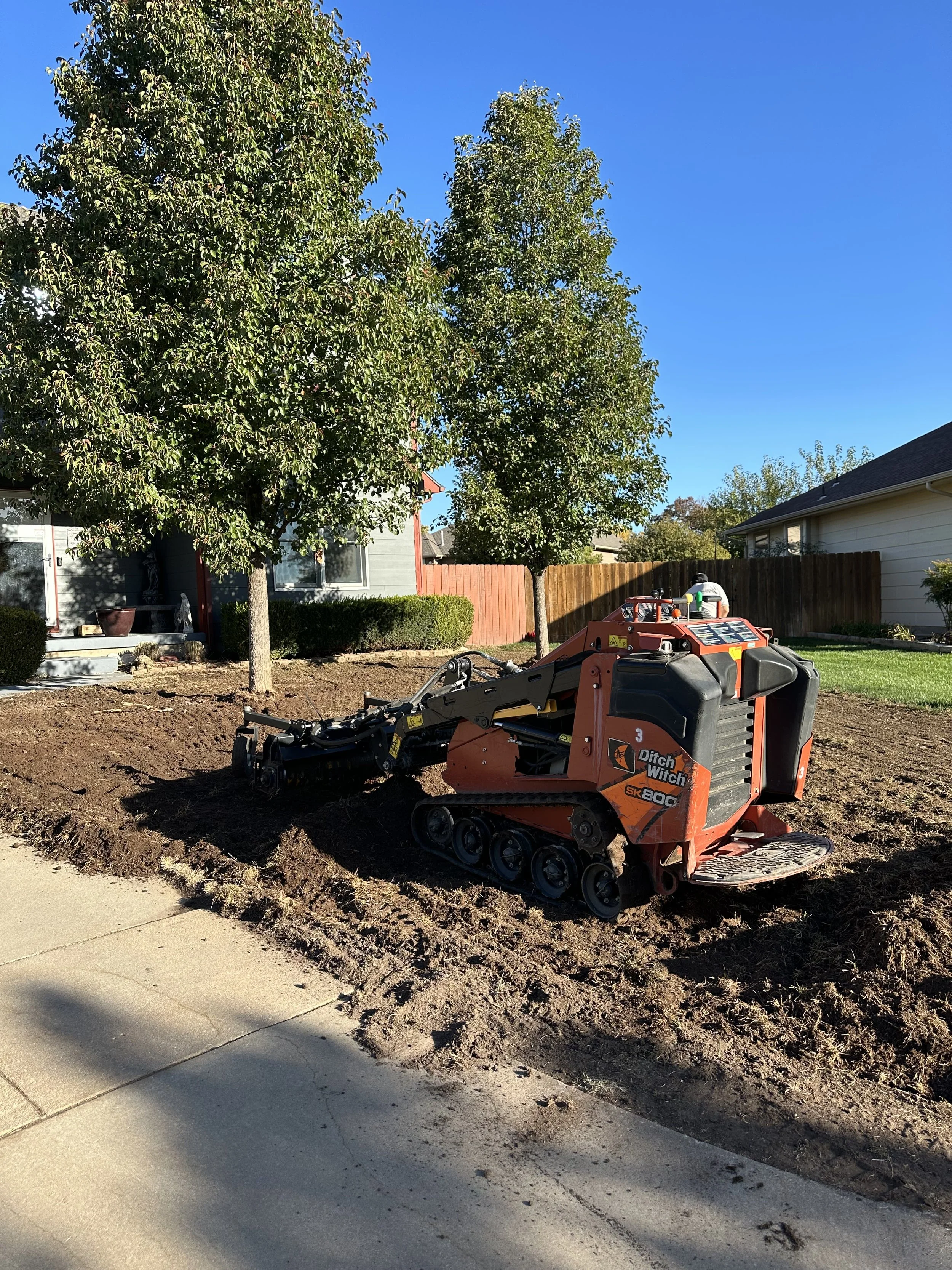 A small orange tracked landscaping machine working on a lawn, with trees and houses in the background, under a clear blue sky.