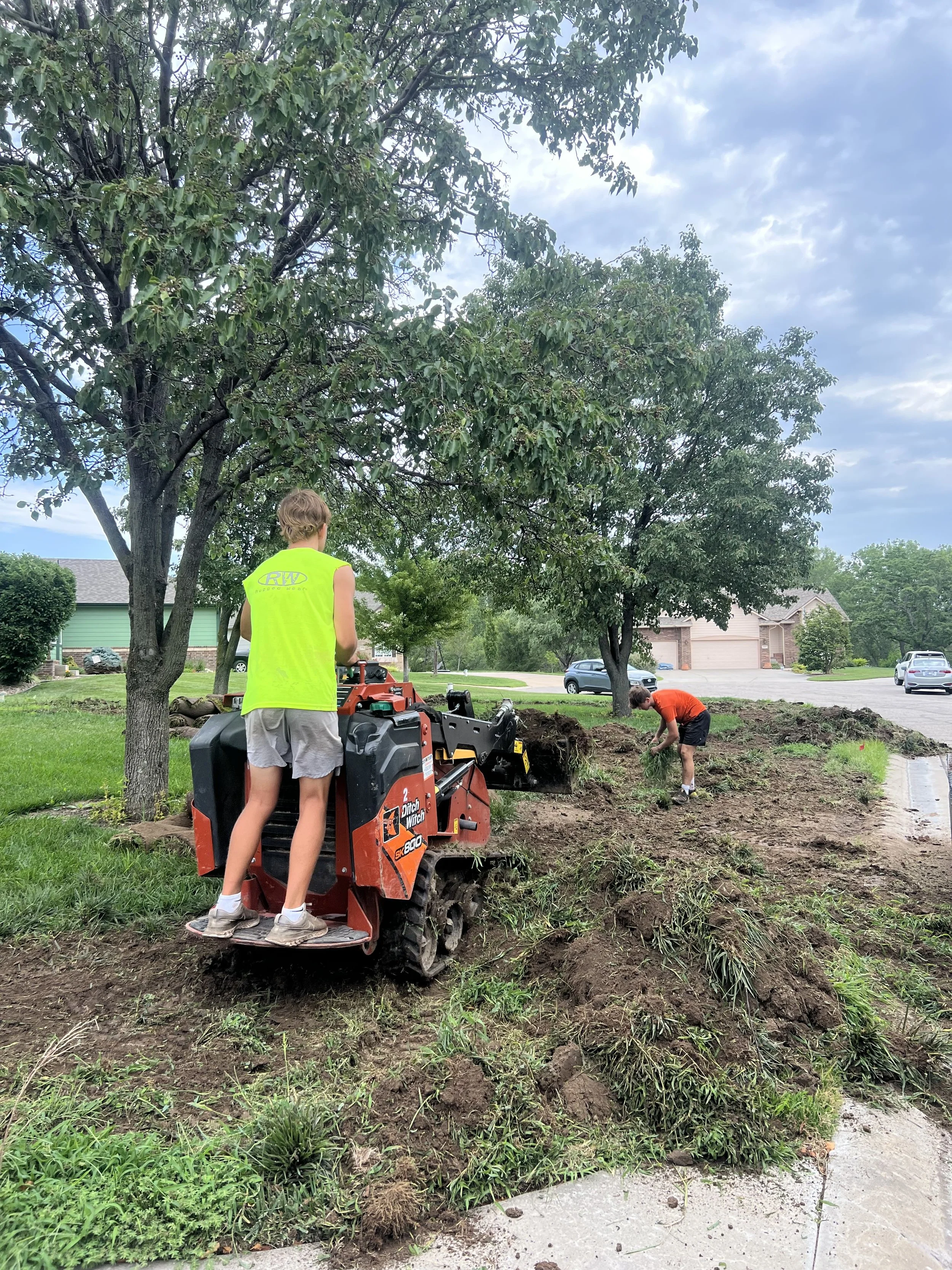 Two children working outdoors on a landscaping project. One child is standing on a mini excavator, wearing a bright yellow sleeveless shirt and shorts, while the other child in an orange shirt is digging in the soil with a tool. There are trees, gras