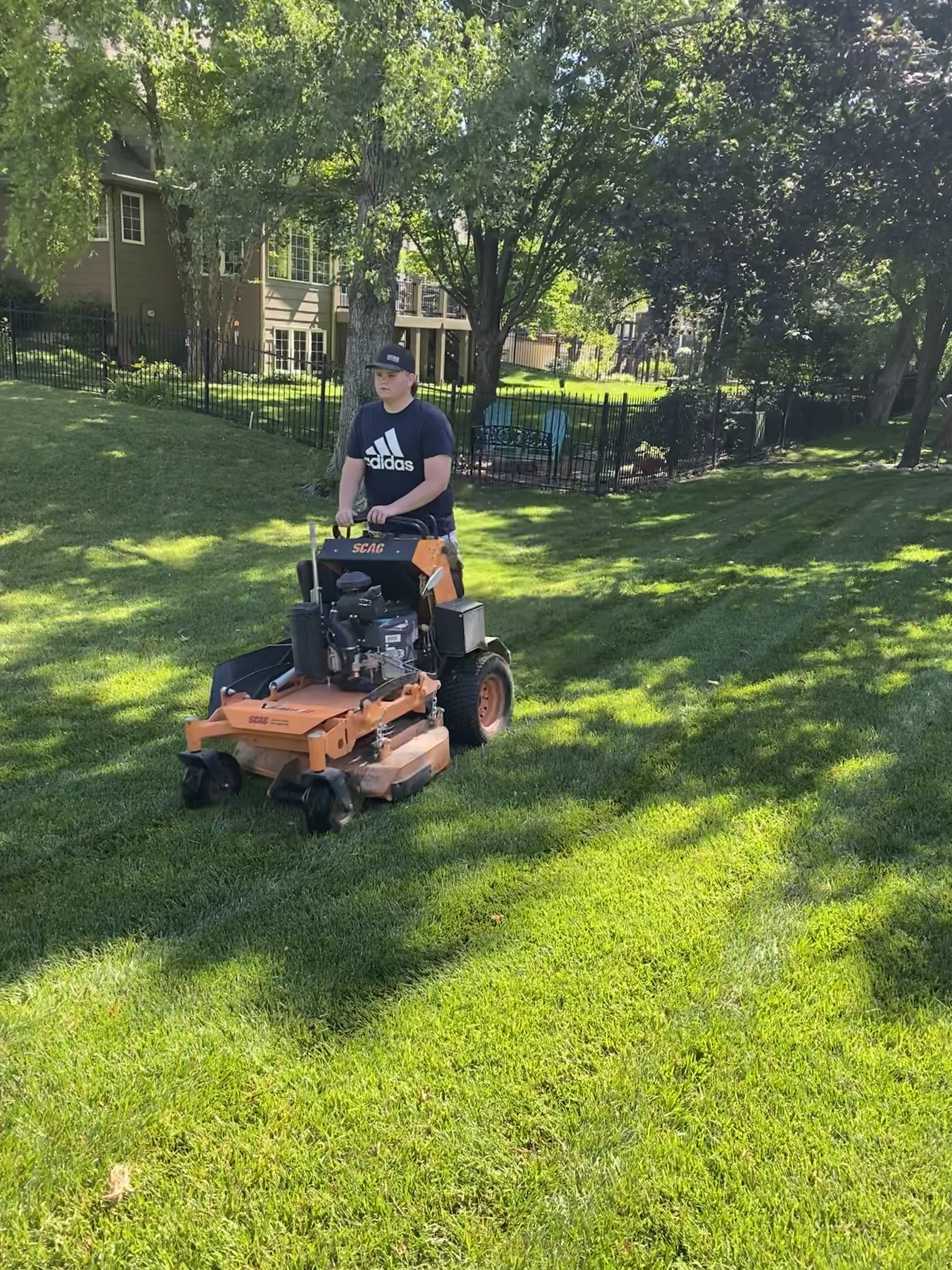 A young man is mowing the lawn with an orange and black riding lawn mower on a sunny day in a residential backyard with green grass, trees, and a dark green house in the background.
