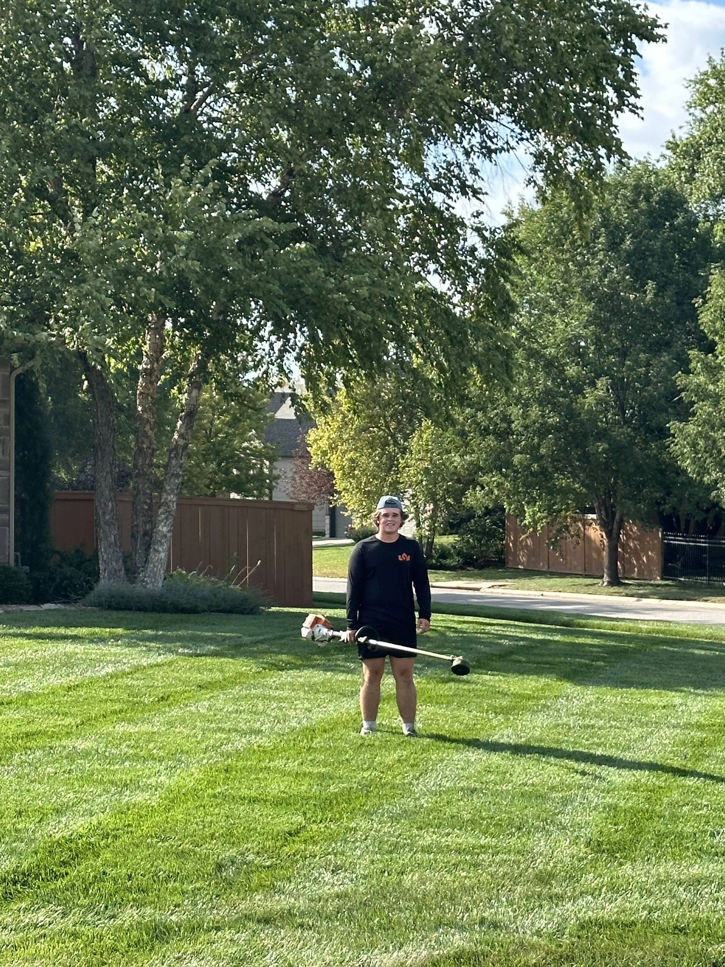 A person stands on a well-maintained grassy yard, holding a string trimmer, wearing a backwards cap, a black long-sleeve shirt, black shorts, and white sneakers, with trees and a wooden fence in the background on a sunny day.