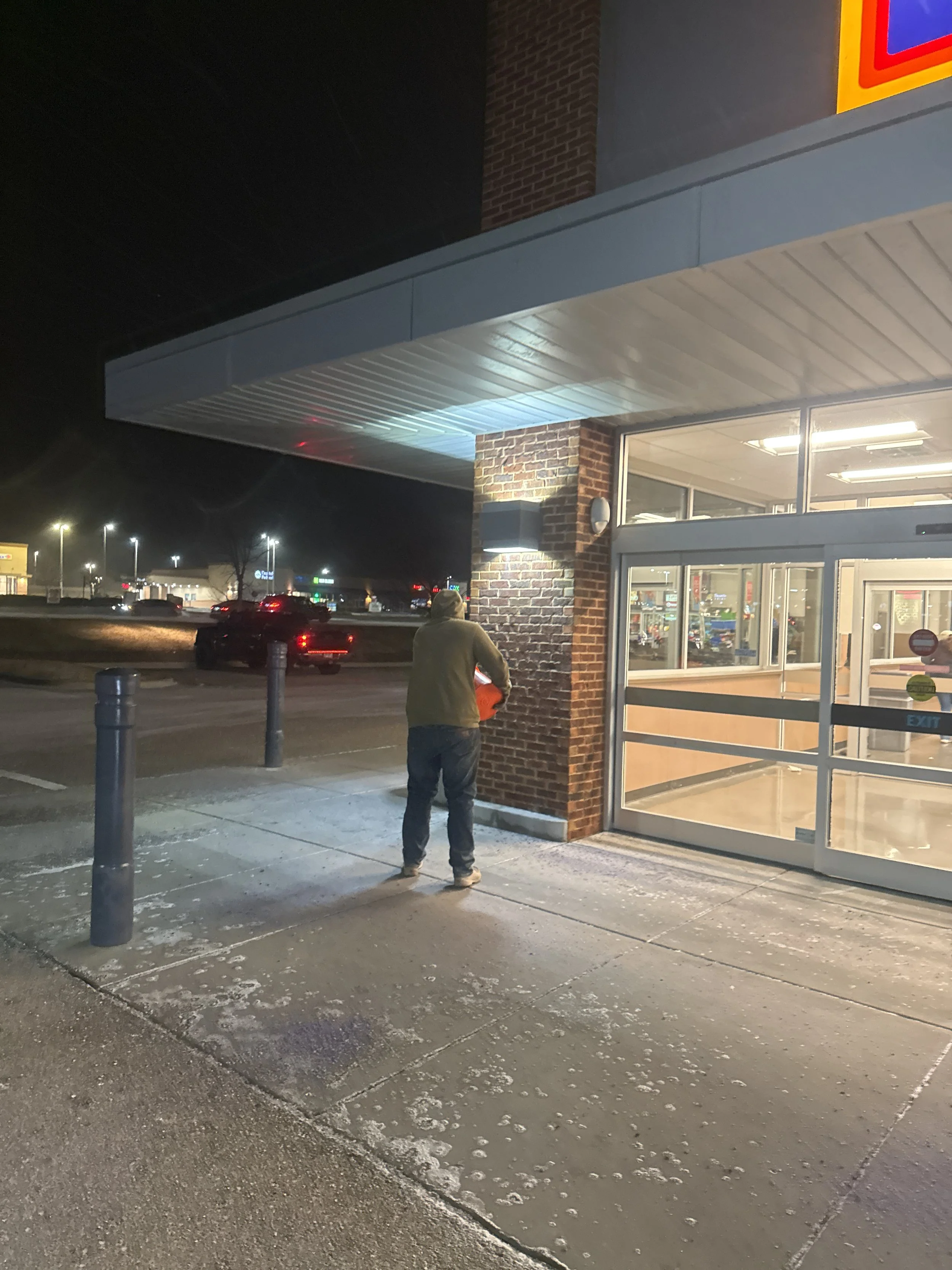 A person standing outside a grocery store at night, holding an orange shopping basket near the entrance.