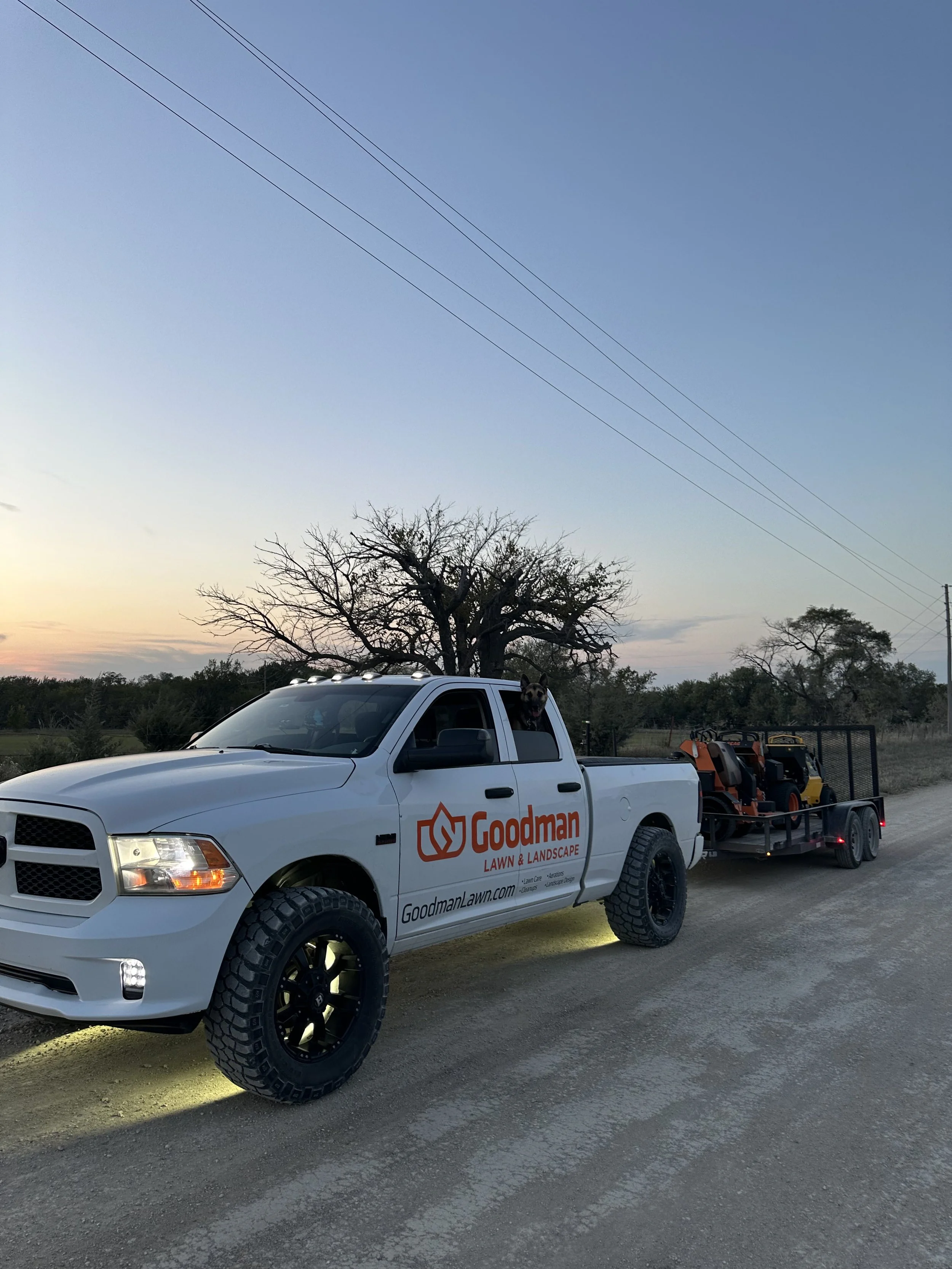 A white pickup truck with black wheels and off-road tires parked on a dirt road at sunset. The truck has the logo and name 'Goodman Lawn & Landscape' on the side, with a dog sticking its head out of the passenger side window. Behind the truck, a trai