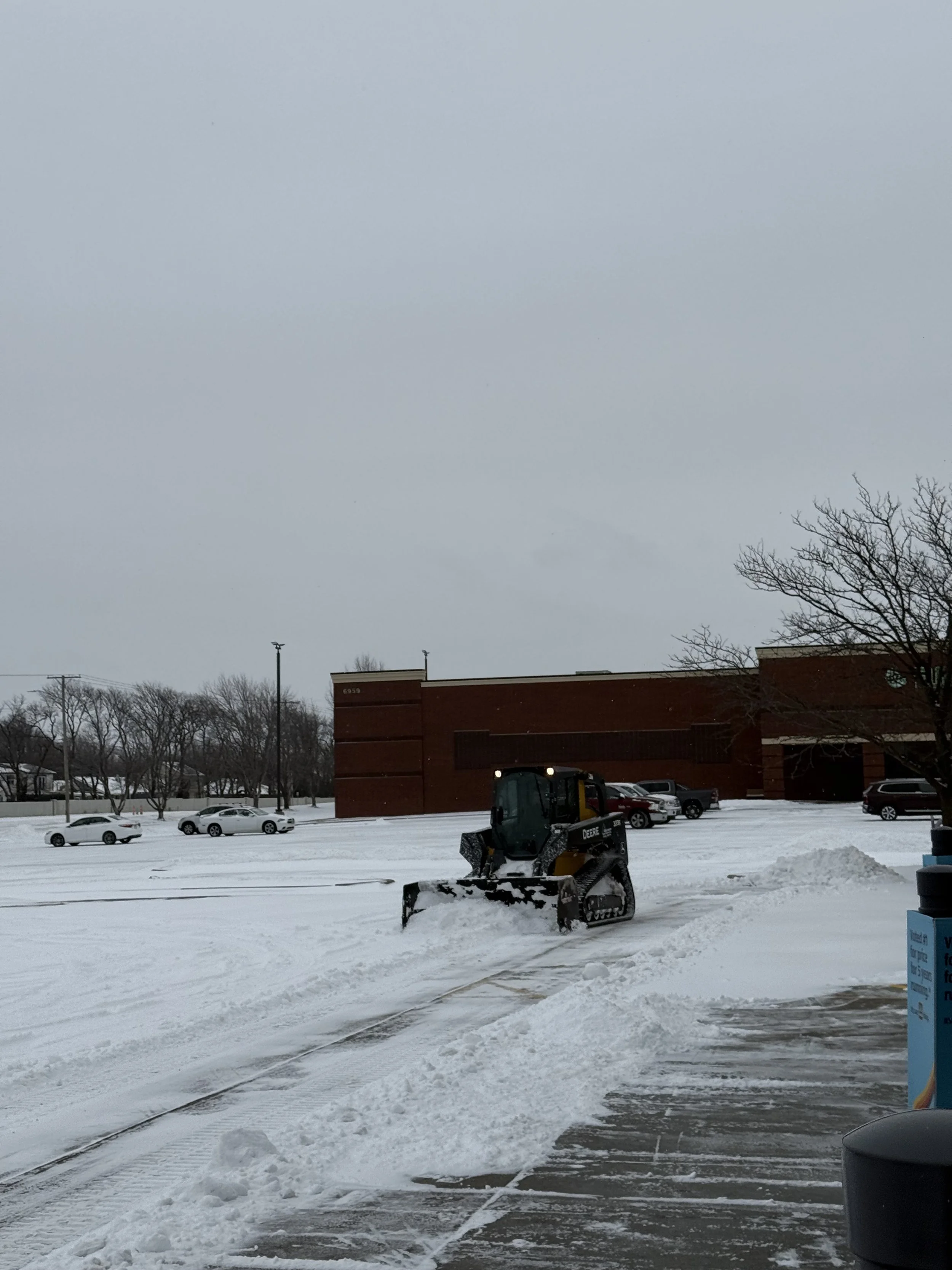 A snow-covered parking lot with a small bulldozer clearing snow, and parked cars in the background. There is a brick building to the right and a leafless tree nearby. The sky is overcast.