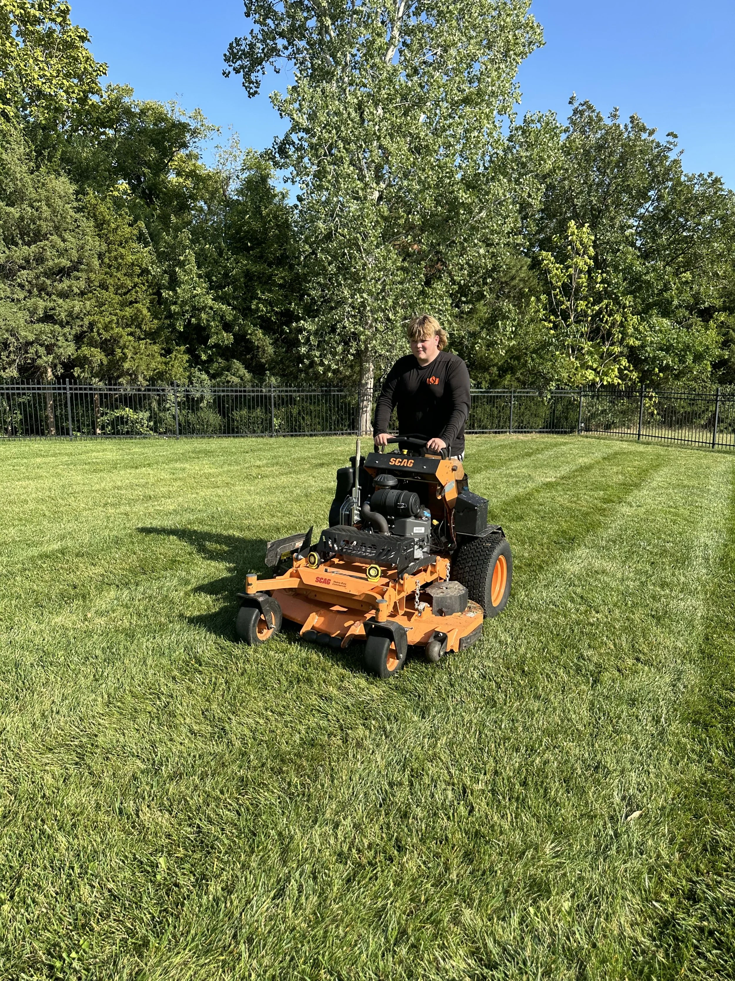 A person operating a large orange and black lawn mower on a well-maintained grassy field with trees and a black metal fence in the background on a sunny day.