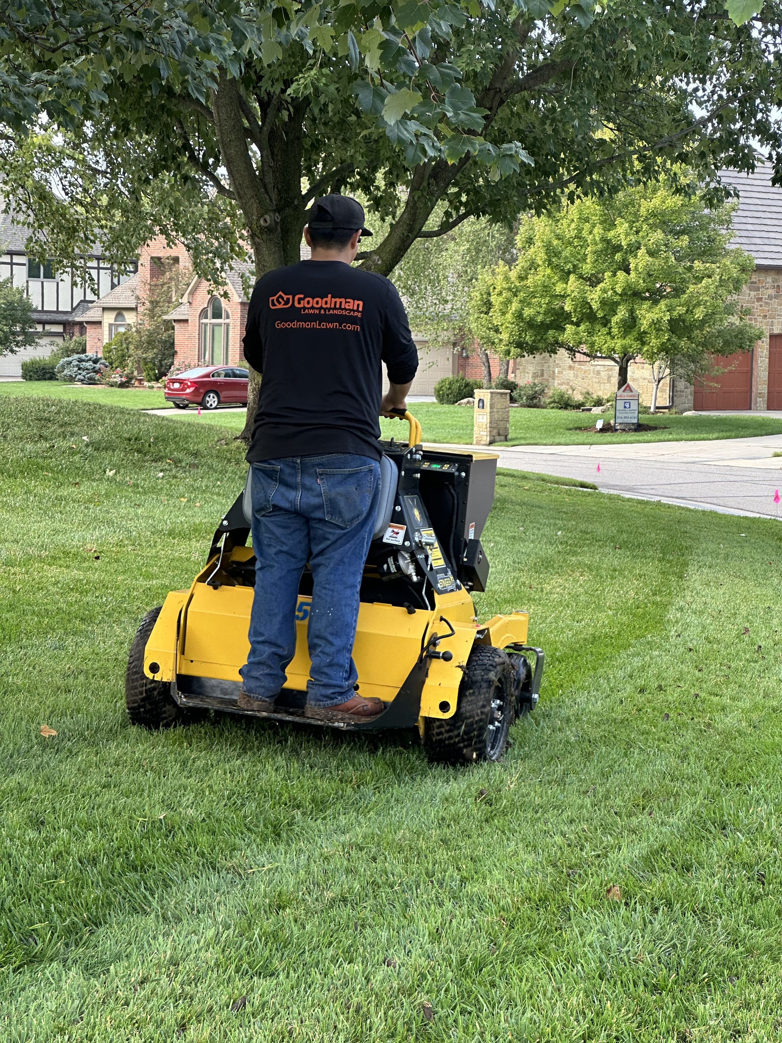 A man wearing a black Goodman Lawn & Landscape shirt operating a yellow lawn mower on a grassy yard in a residential neighborhood.