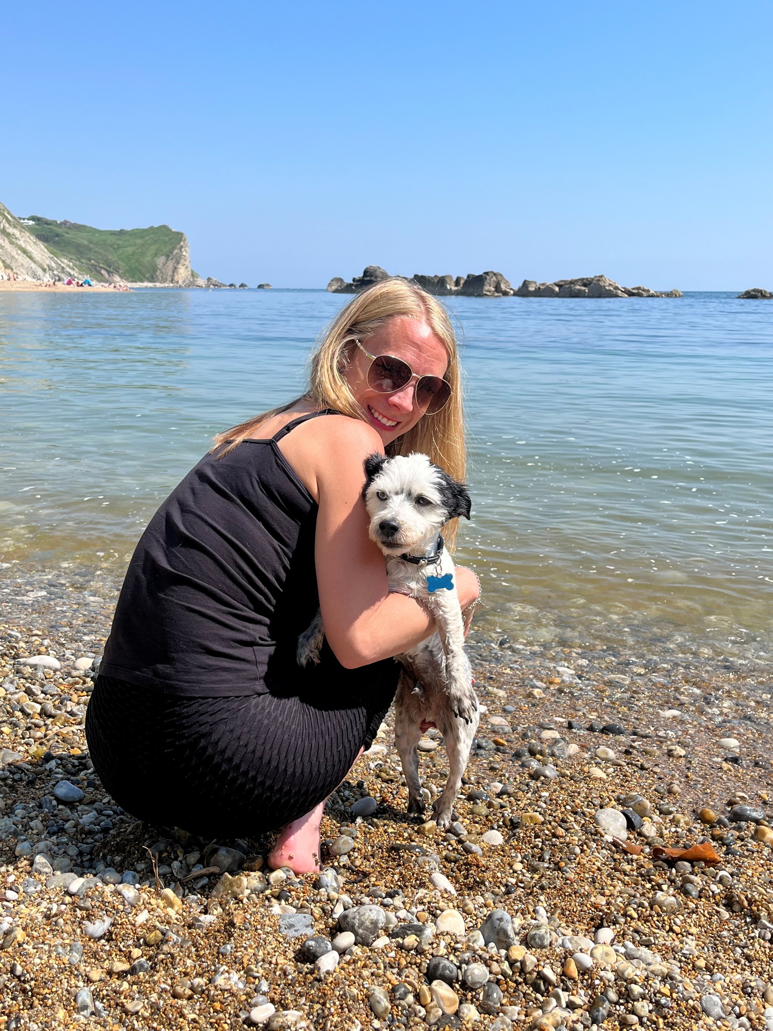 A woman with long blonde hair wearing sunglasses, kneeling on a rocky beach holding a black and white puppy near the water. In the background, the sea, rocks, and cliffs are visible under a clear blue sky.