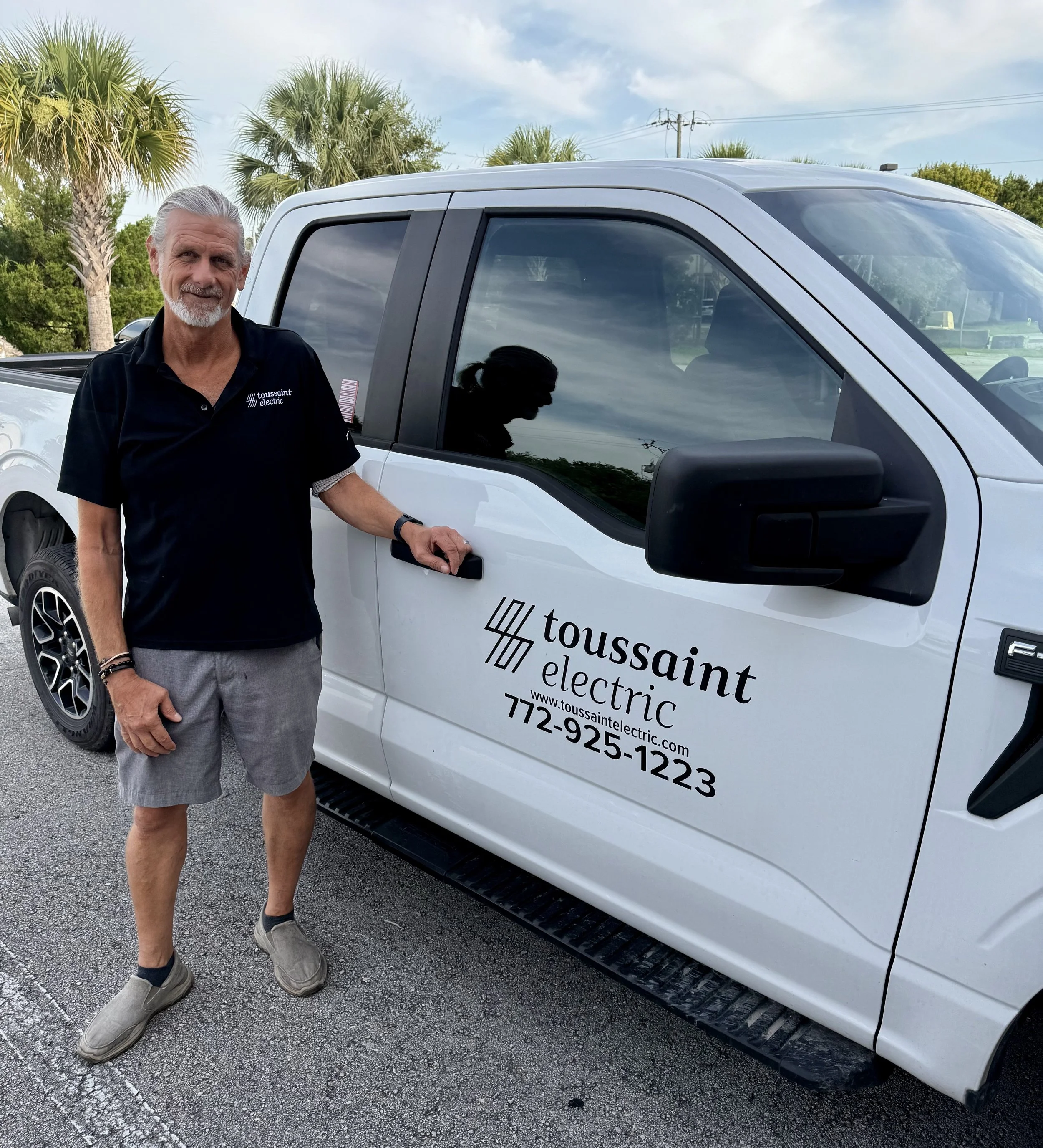A middle-aged man with gray hair and a beard standing outdoors next to a white electric truck with 'Toussaint Electric' branding on the door, palm trees in the background, wearing a black polo shirt and gray shorts.