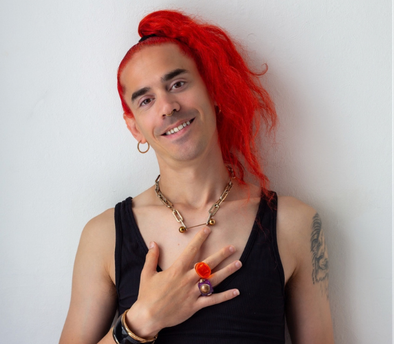 A white person with a soft smile and long orange hair, in the center of the image, holding their hand in the middle of their chest