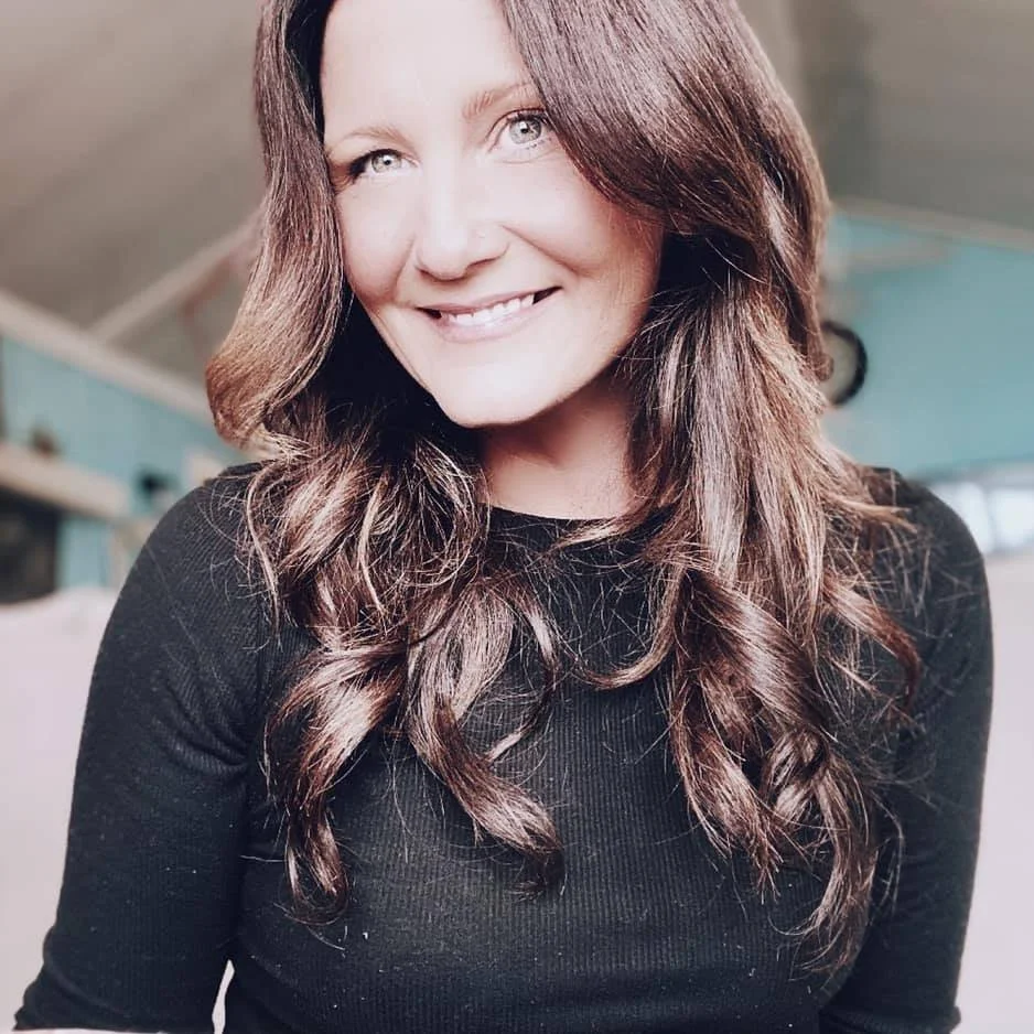 Woman smiling, with long brown hair, wearing a black top indoors.