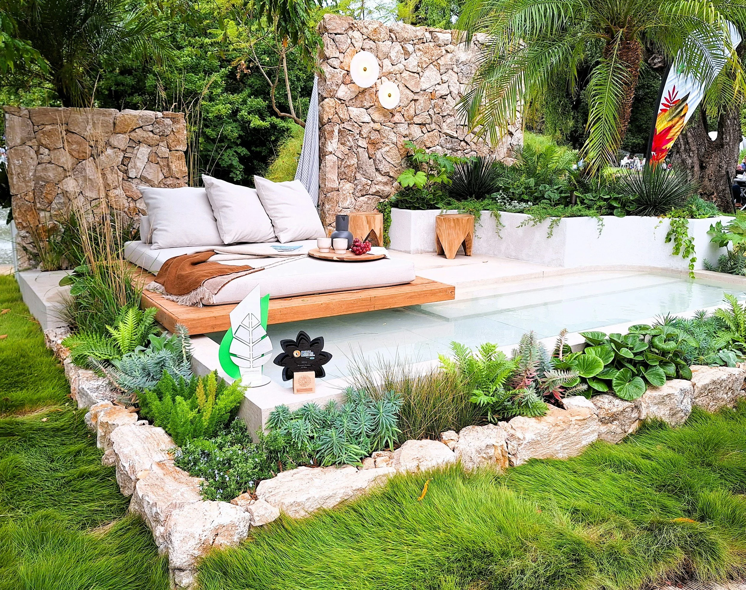Outdoor seating area with a cushioned bench, throw blanket, and decorative vases on a tray, surrounded by lush green plants and a stone wall, set in a garden.