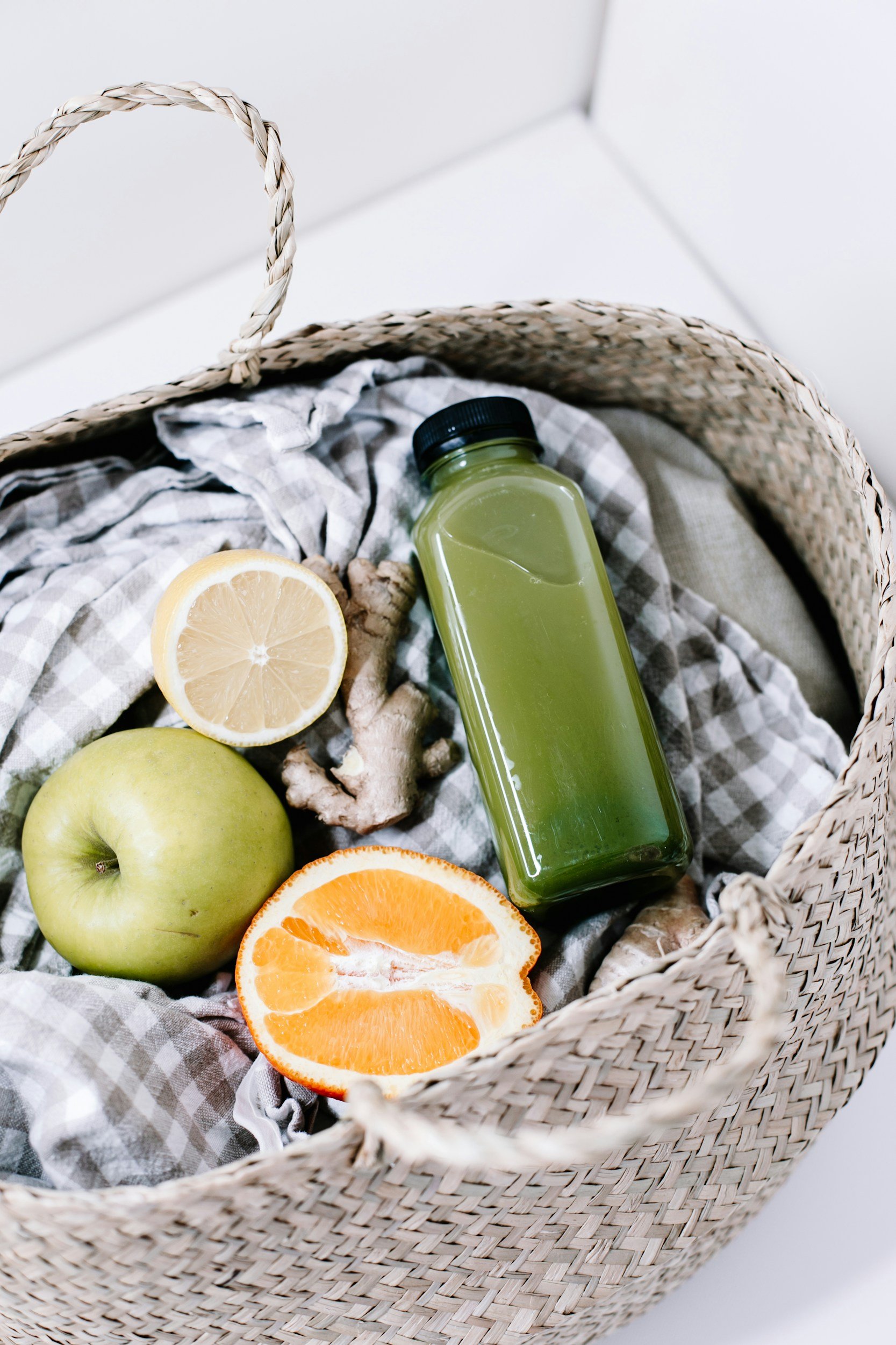 A basket containing a green apple, a lemon half, an orange half, a piece of ginger, a bottle of green juice, and a gray checkered cloth.