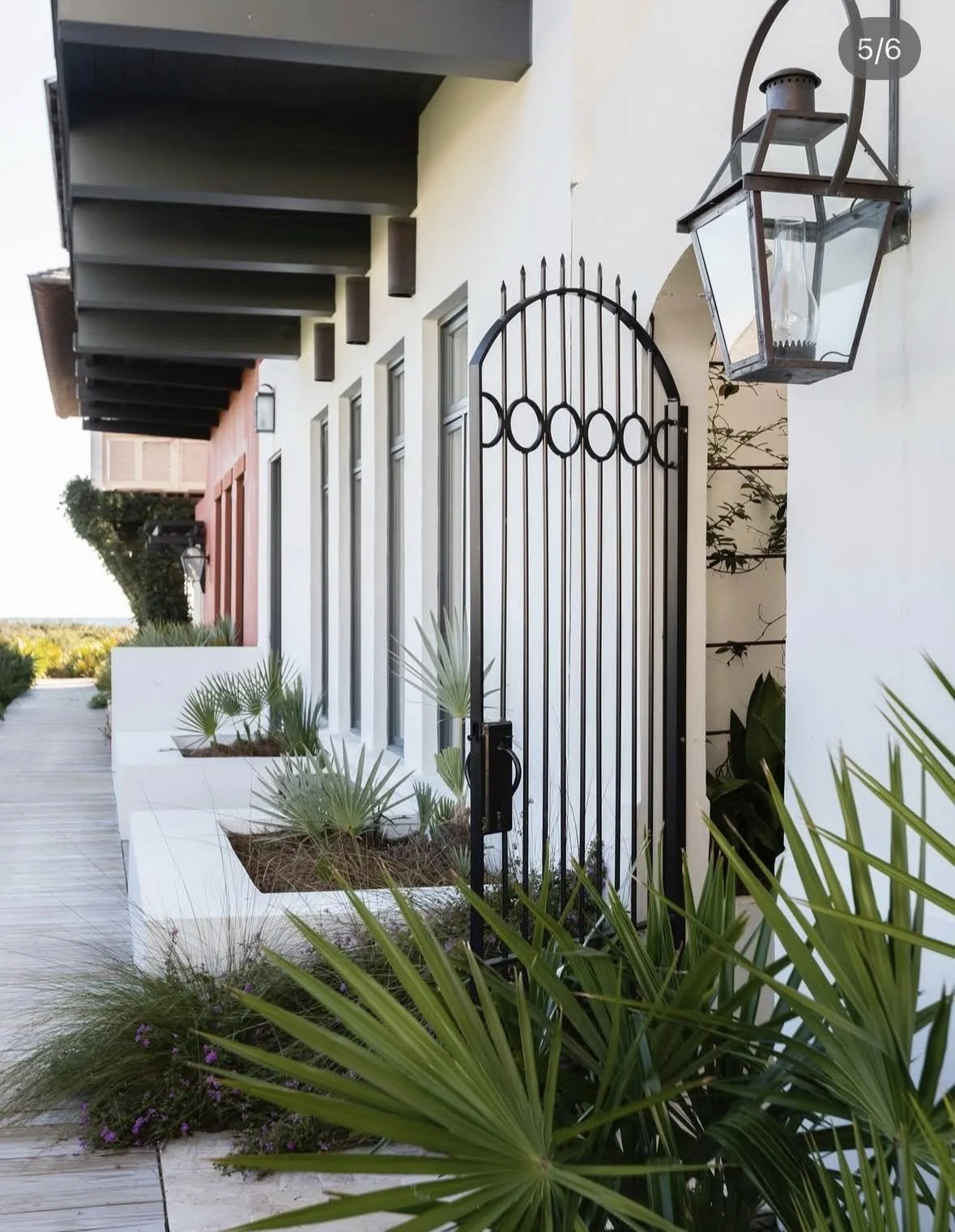 Exterior view of a building with modern design, featuring black metal gate, large lantern light fixture, plants in white planters, and a wooden deck walkway.