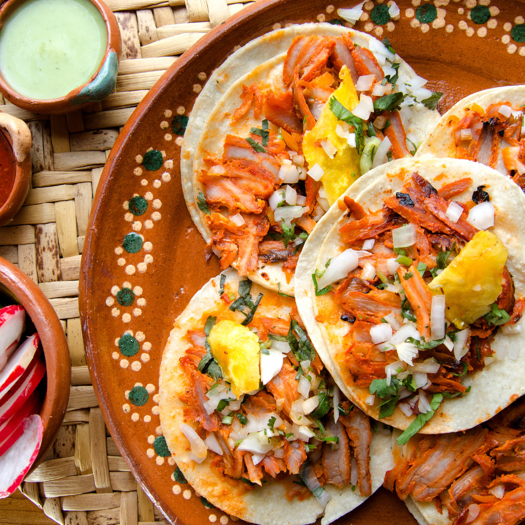 Close-up of three tacos with shredded meat, chopped onions, cilantro, and a piece of yellow grilled cheese, served on a decorative brown plate.