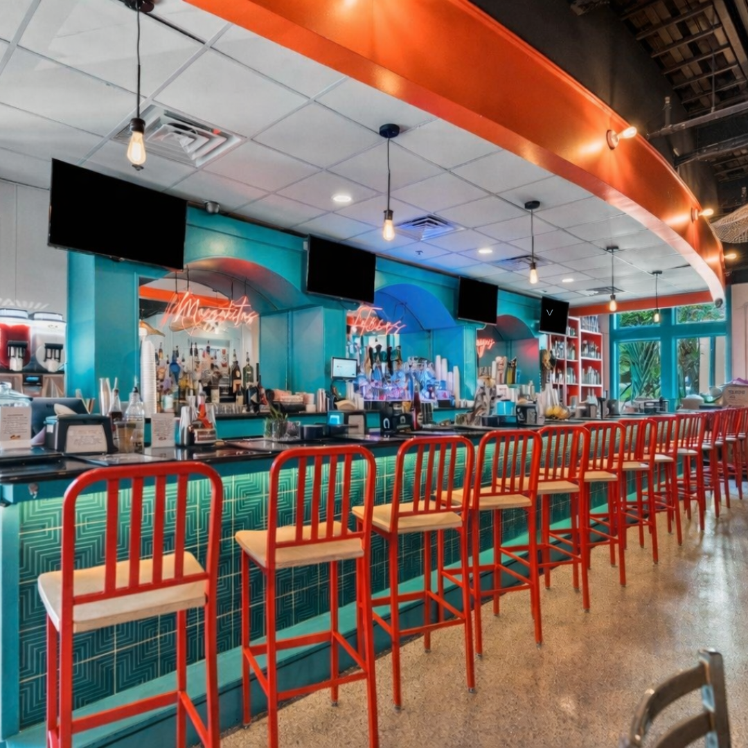 A colorful cafe or bar with a turquoise counter, red bar stools, and a modern design. Several screens hang above the counter, and neon signs glow in the background. There are shelves with bottles and drink dispensers behind the counter.