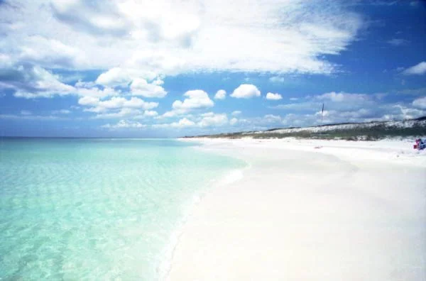 A clear, turquoise ocean meets a white sandy beach under a partly cloudy sky.