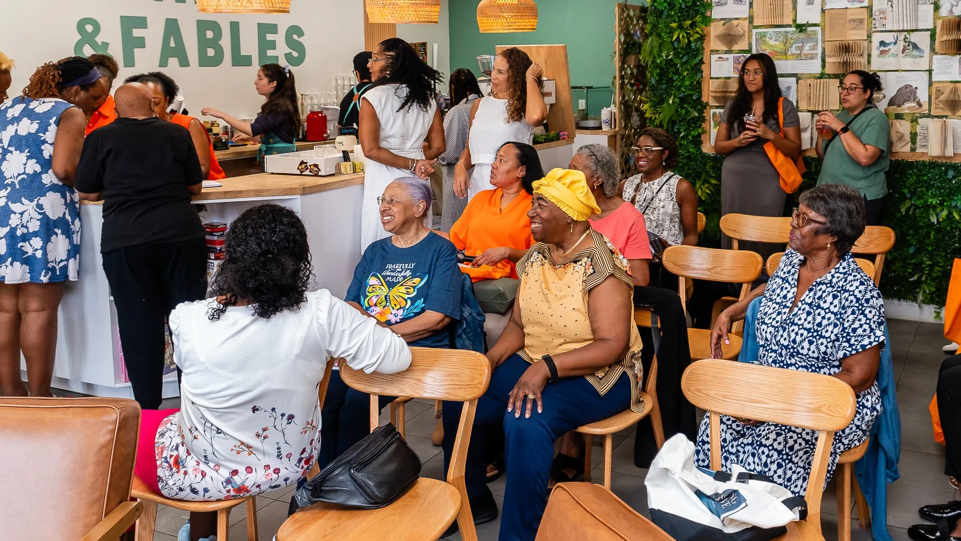 A group of women gathered at a cafe or community space, engaging in conversations and enjoying each other's company. Some are sitting on chairs, smiling and laughing, while others are standing and talking near a counter with a sign that says '& FABLE