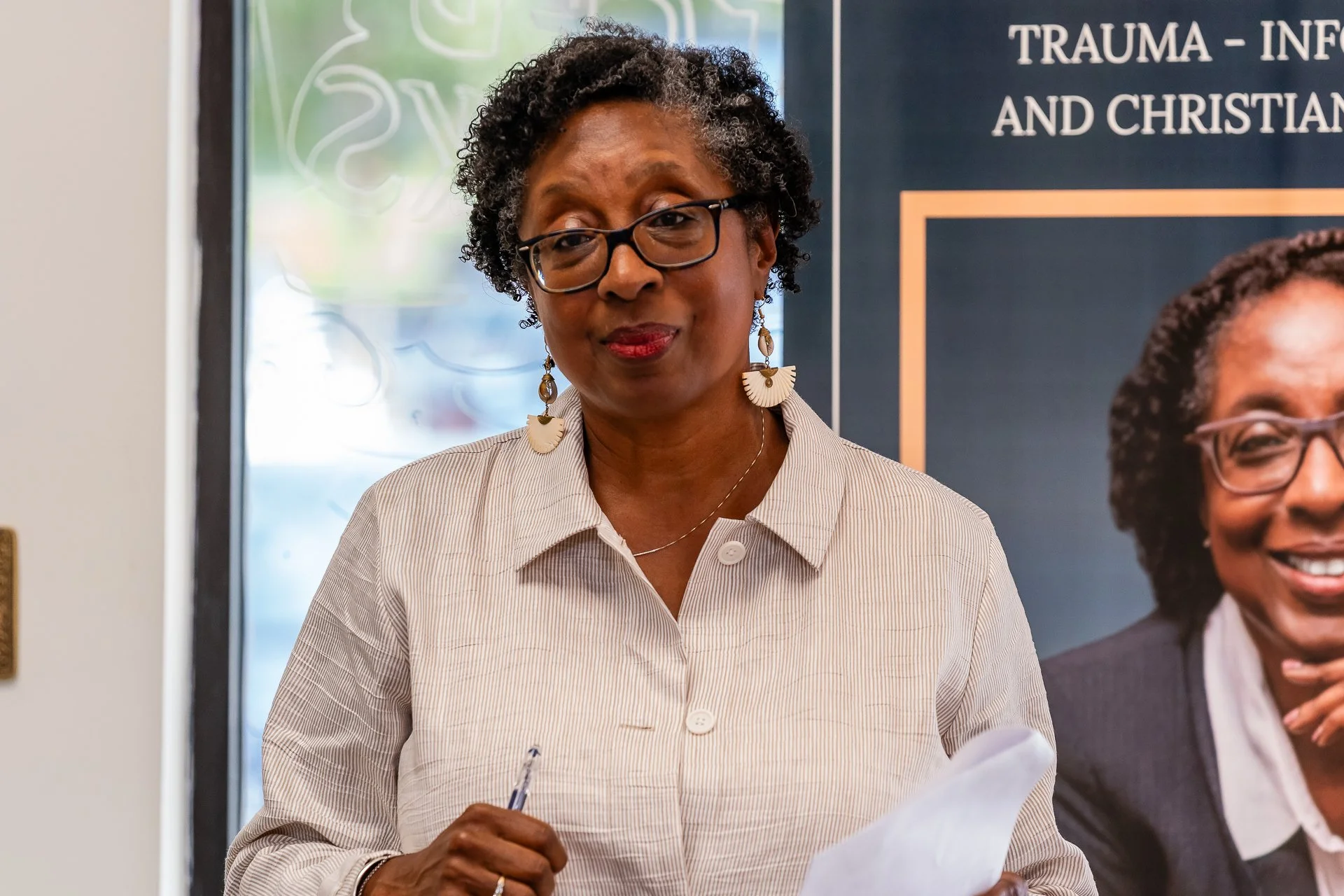 A woman with glasses, curly hair, and wearing a beige striped shirt, standing and holding a pen and paper, in front of a sign that says "Trauma-Informed and Christian"