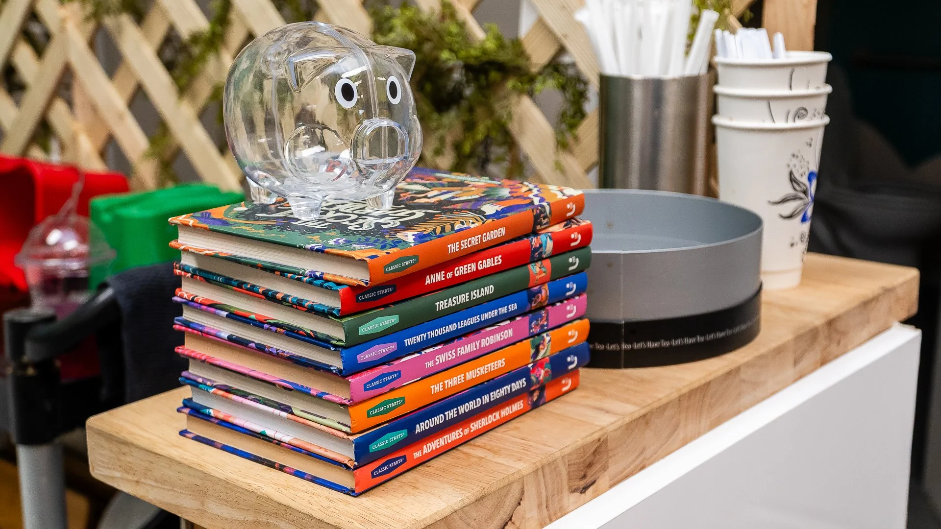Stack of colorful paperback books on a wooden table, with a clear piggy bank on top. In the background, there are white cups, paper straws, and a plant.