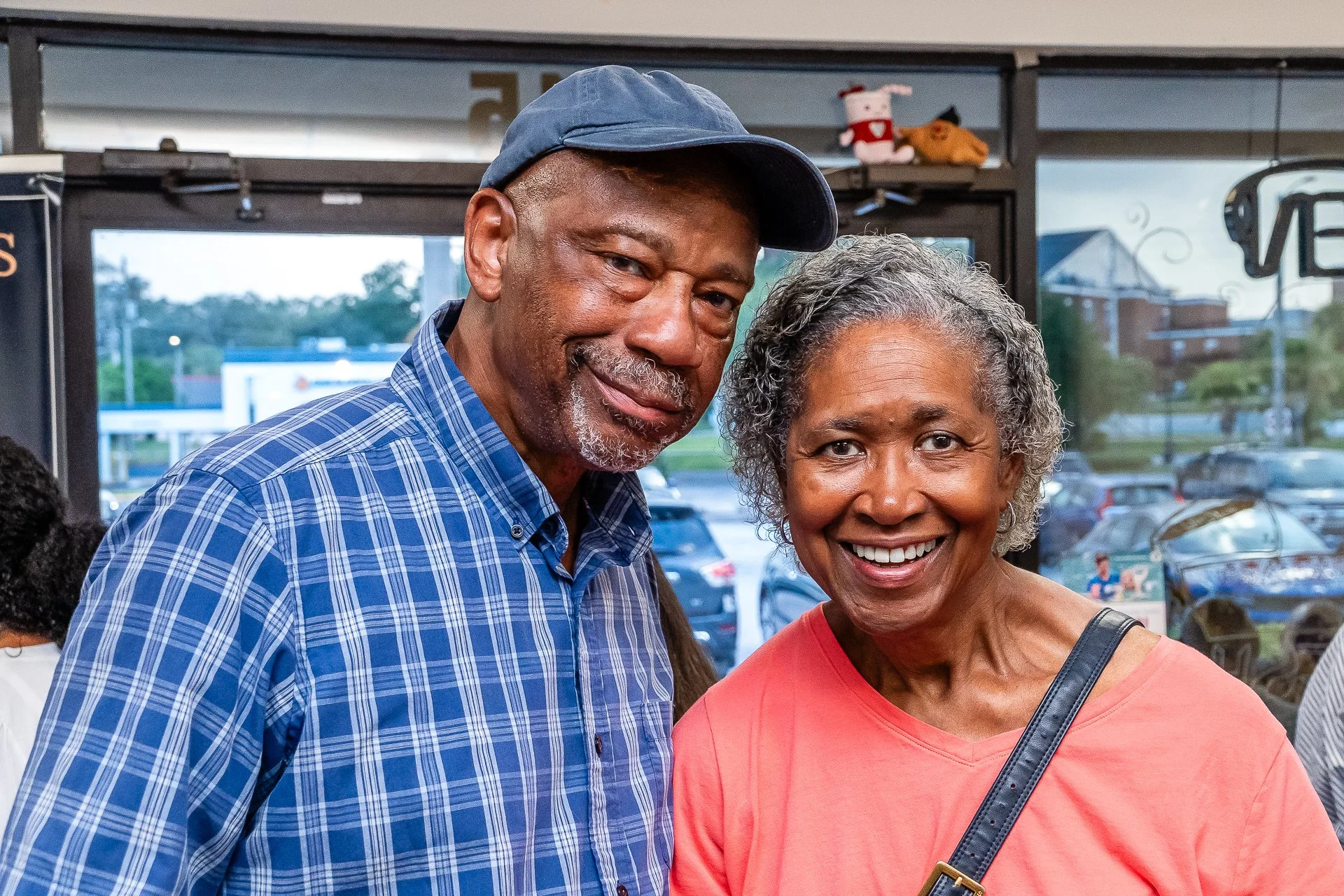 A smiling African American woman in a pink shirt standing next to a smiling African American man wearing a blue plaid shirt and a blue cap inside a business or restaurant with an outdoor parking lot visible through the windows.