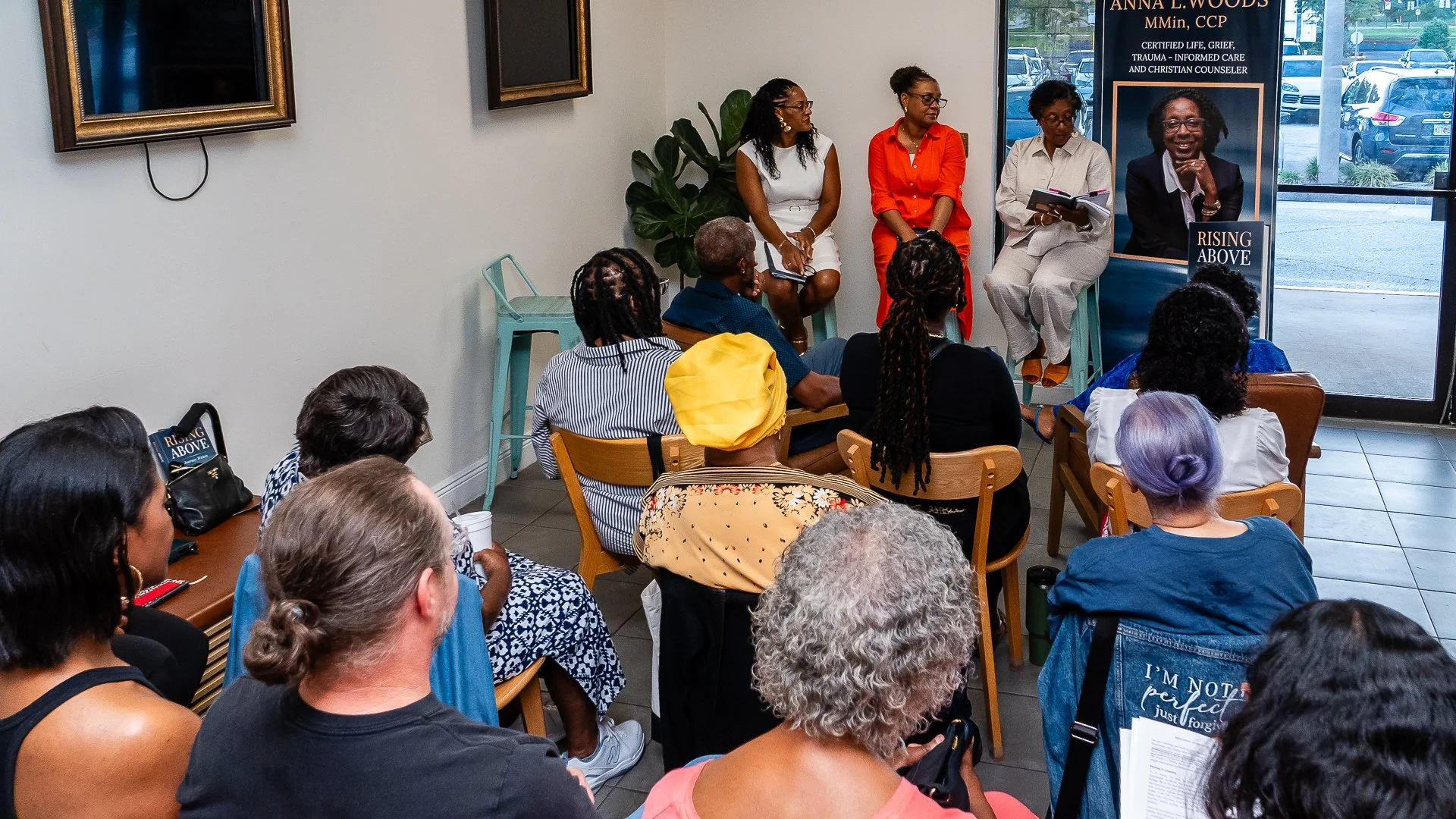 A panel discussion with five women seated in front of an audience in a room with blue chairs, large windows, and artwork on the wall. One panelist is reading from a book, and a poster behind her displays her photo and credentials.