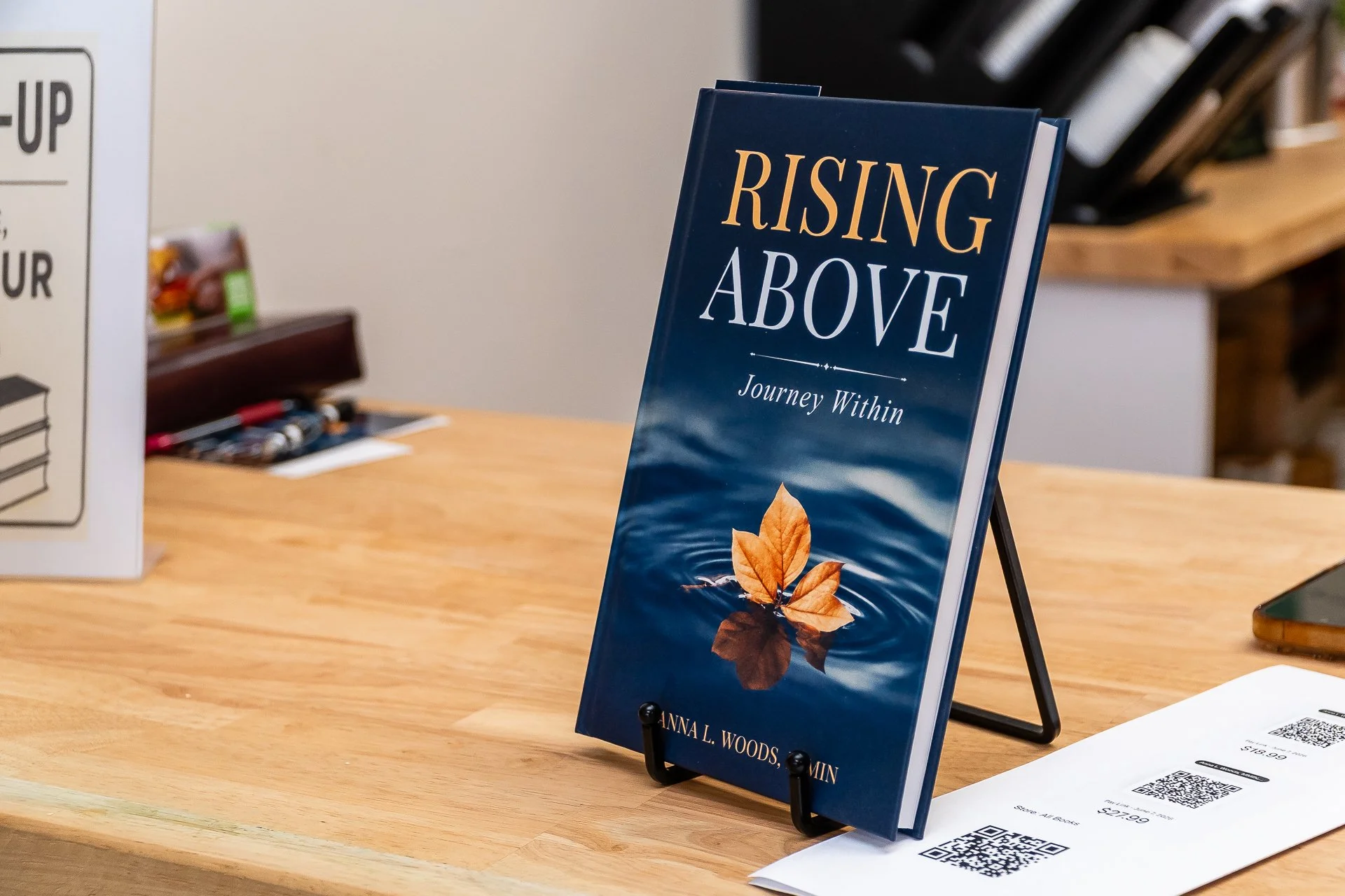 Book titled "Rising Above: Journey Within" by Anna L. Woods Minn displayed on a table, with QR code and receipt nearby.