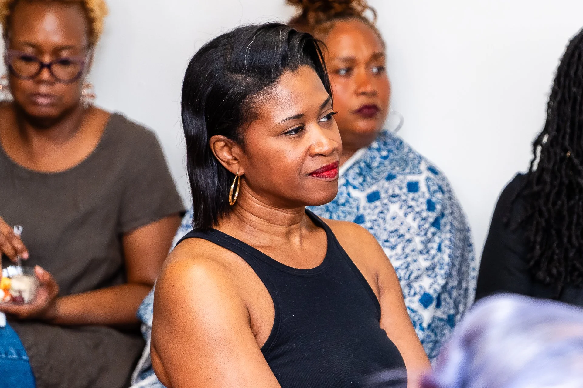 A group of women sitting together at an indoor event, with one woman in the foreground wearing a black sleeveless top and gold hoop earrings, looking attentively to the side.
