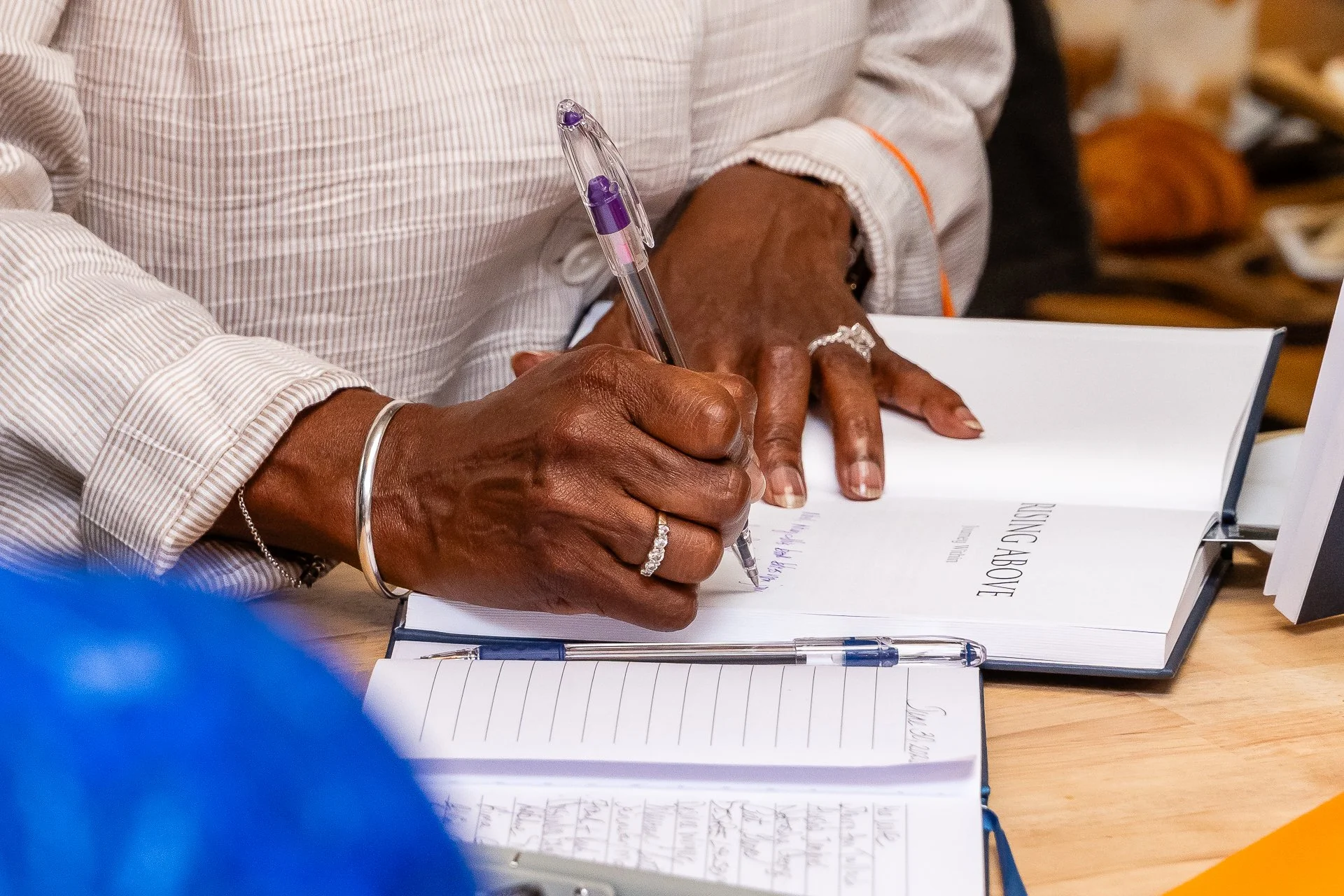 A person with dark skin wearing rings and bracelets signs a book using a purple and clear pen at a table, with a blurred background.