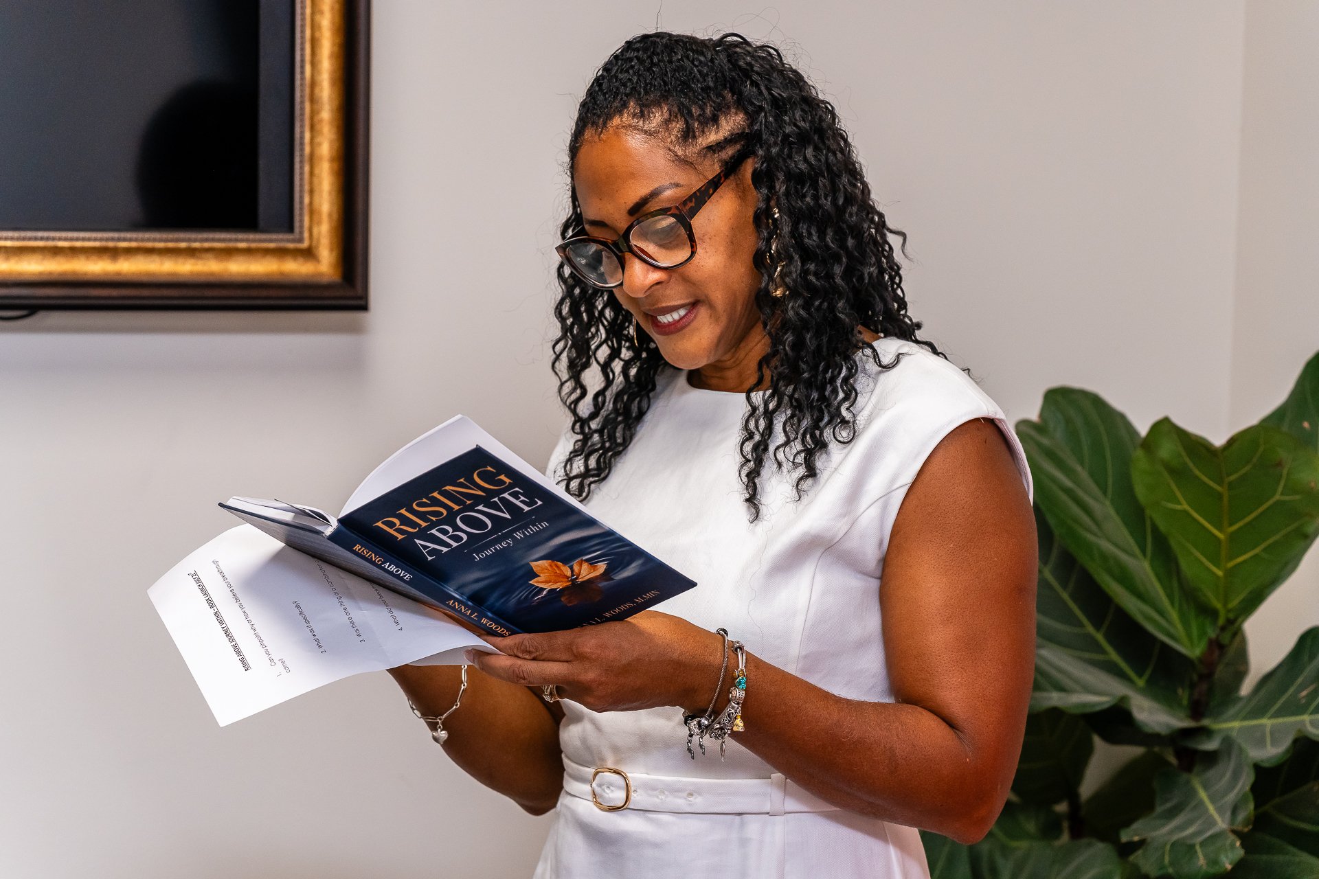 A woman with curly black hair and glasses reading a book titled 'Rising Above' in an indoor setting, with a large green plant behind her and framed artwork on the wall.
