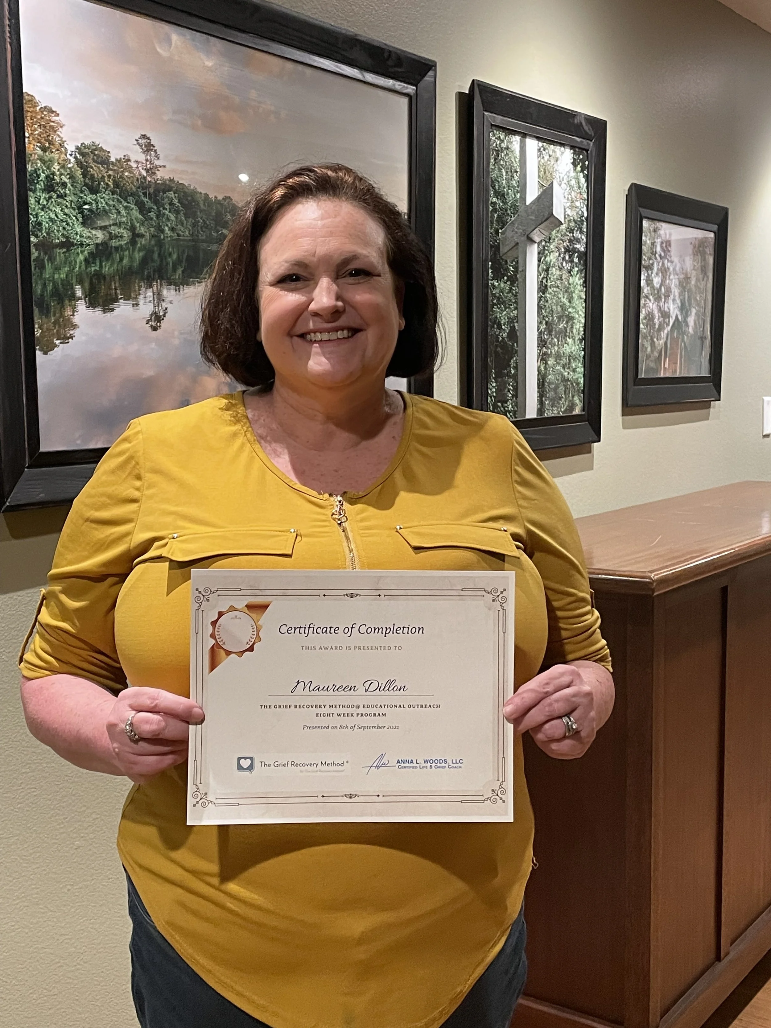 Woman in yellow shirt smiling and holding a certificate of completion in front of framed landscape photos on a wall.