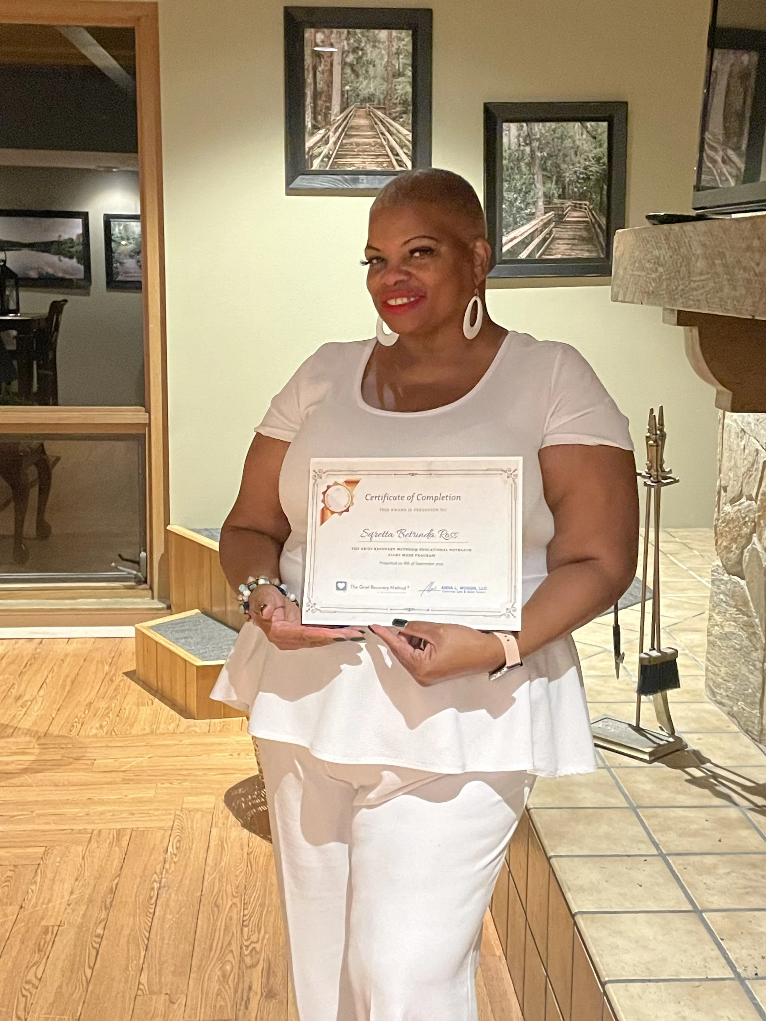 Woman with short hair and large earrings holding a certificate of completion, standing in a room with wood and stone decor and framed pictures of a wooden walkway on the wall.