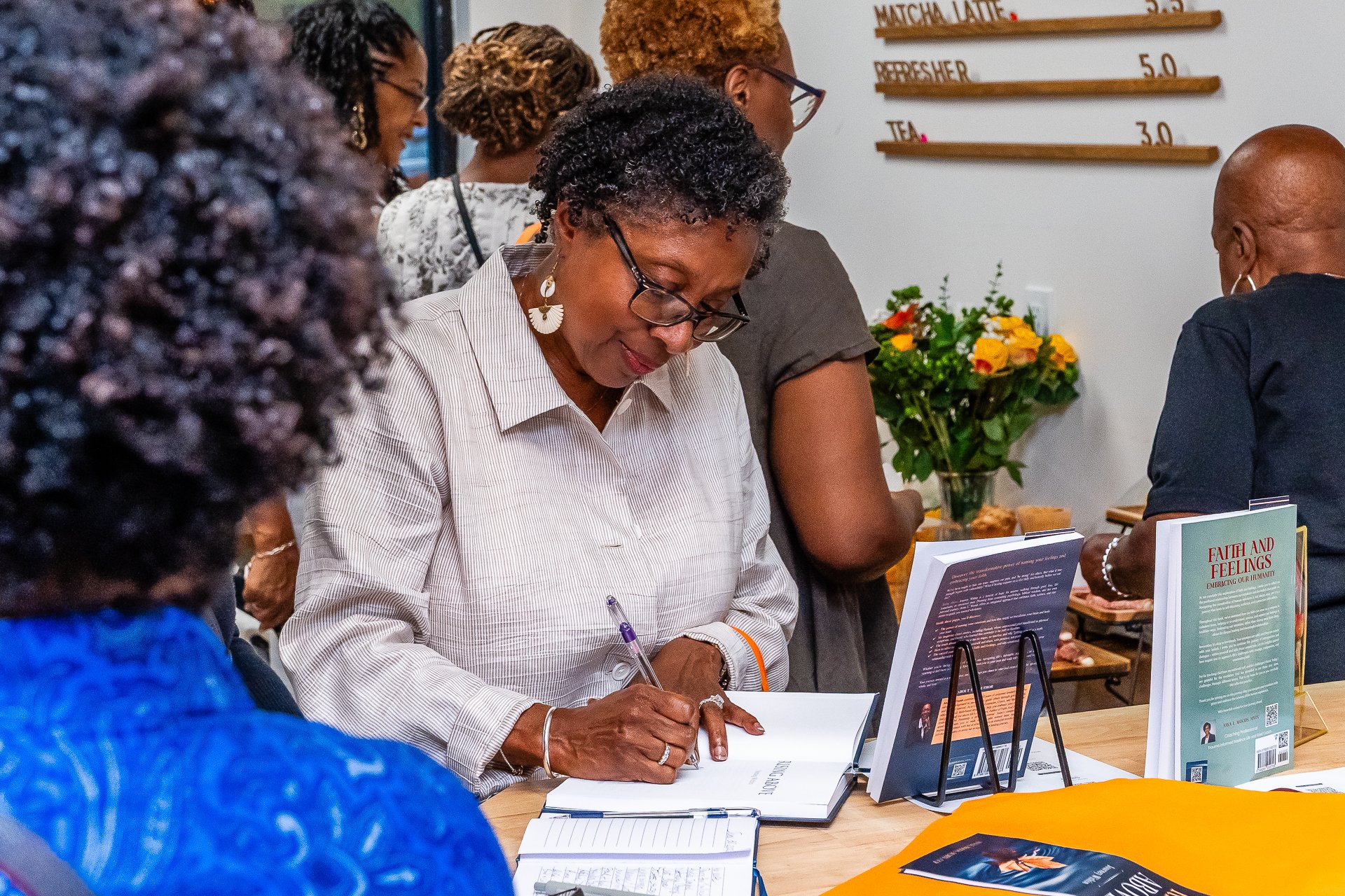 A woman with short curly dark hair, glasses, and earrings is signing a book at a table during an event. Several people surround her, browsing books and engaging in conversation. The table has informational pamphlets, and a book titled 'Faith and Feel