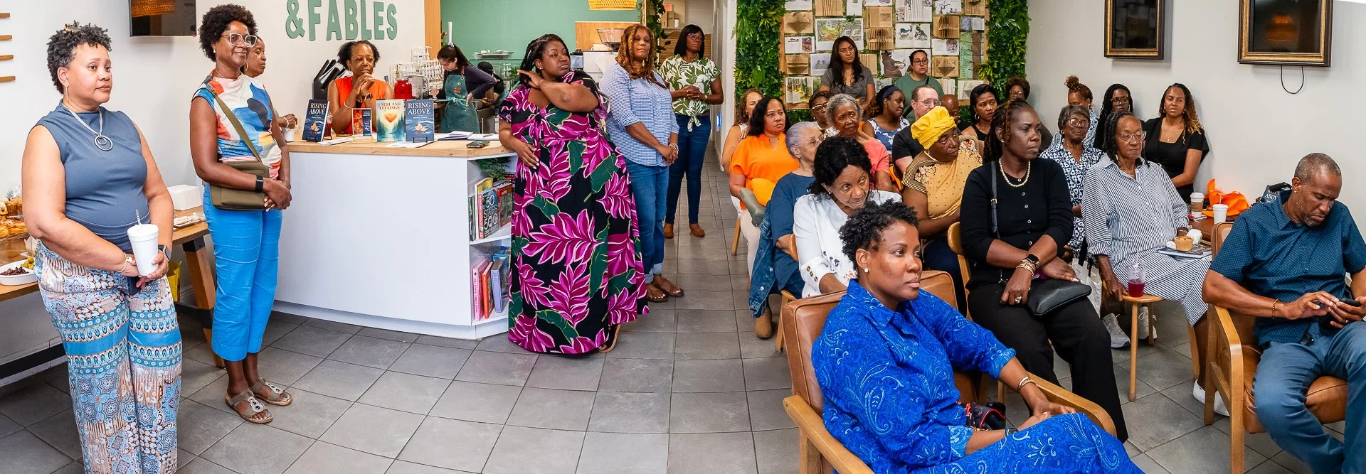 A group of women attending a book reading or discussion event in a cafe or bookstore. Some are standing, others are seated, listening attentively. The space features bookshelves, framed pictures, and a counter with books and drinks.