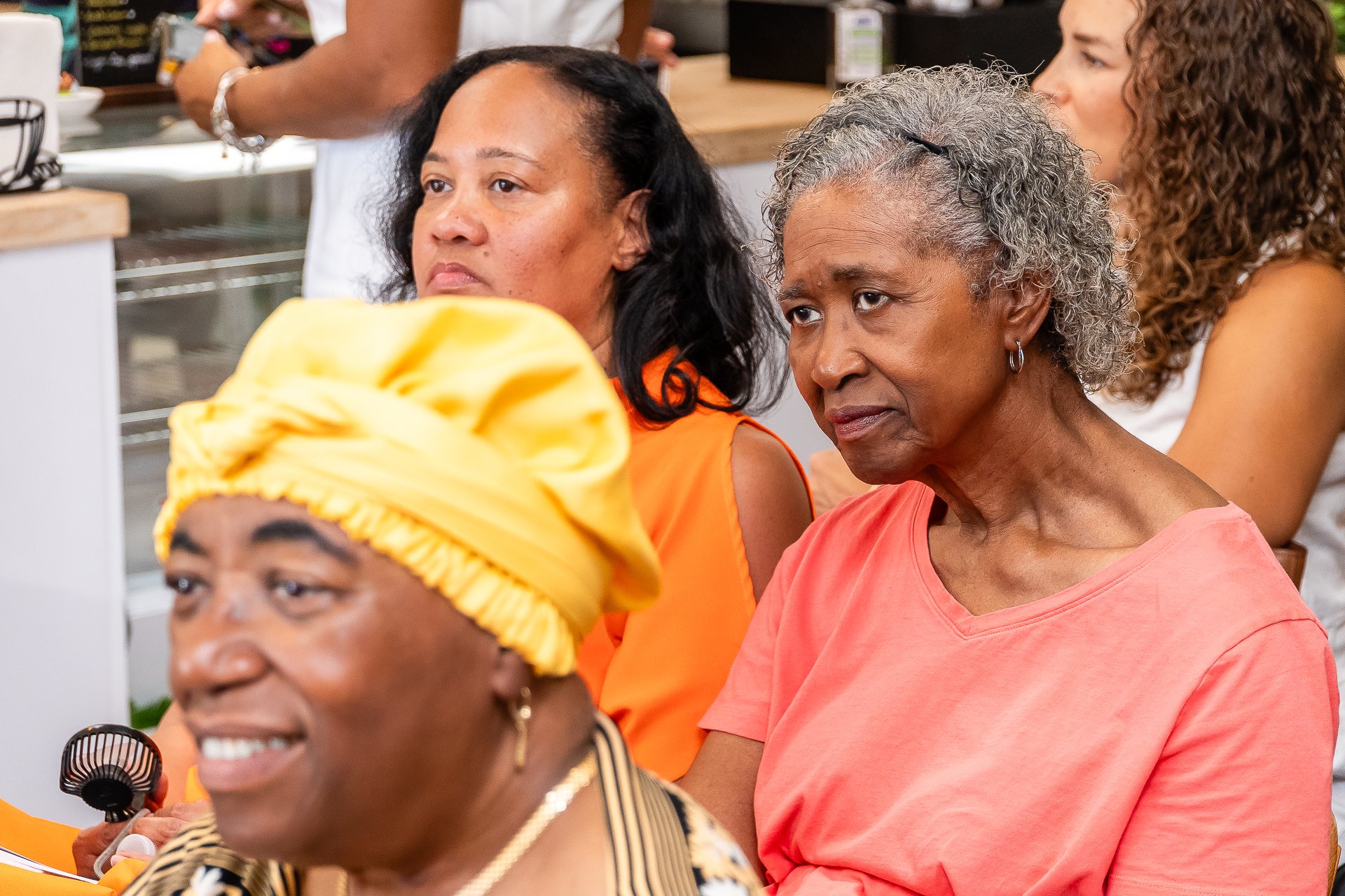 Group of diverse women sitting attentively at an indoor event. One woman in the foreground wearing a yellow headscarf, another in the middle with curly gray hair and orange shirt, and others behind them listening.