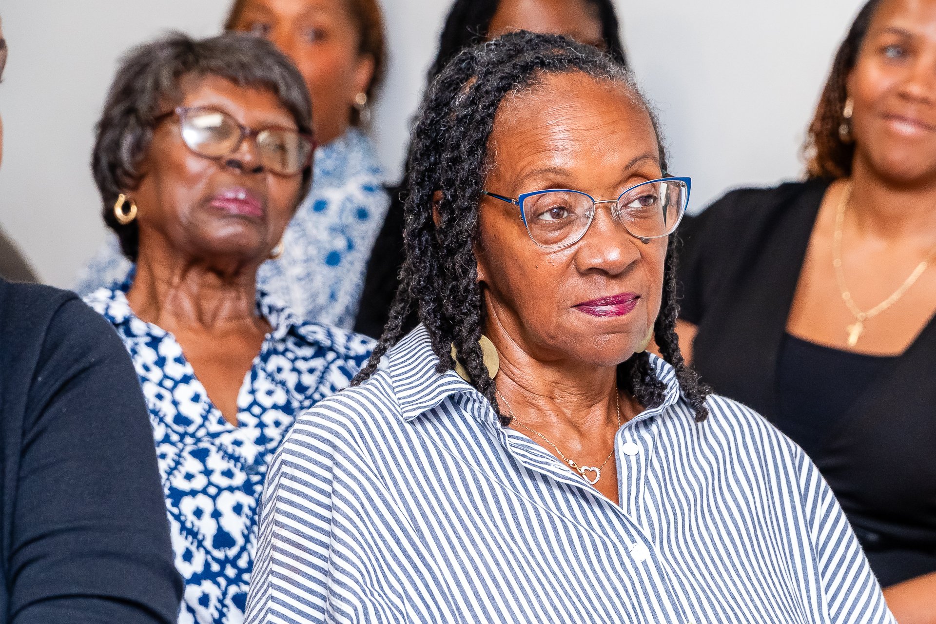 A group of diverse women attending a meeting or event, dressed in business casual attire, with one woman in the foreground wearing a striped shirt and glasses, and others visible in the background.