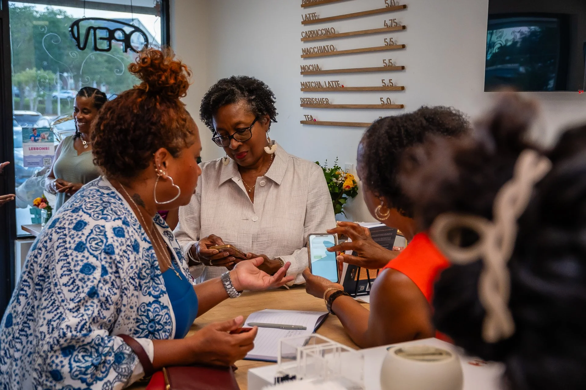 A group of women gathered around a counter inside a cafe, engaging with their phones. The cafe menu is visible on the wall with options like latte, cappuccino, macchiato, and matcha latte. The scene is lively and social, with a woman smiling in the b
